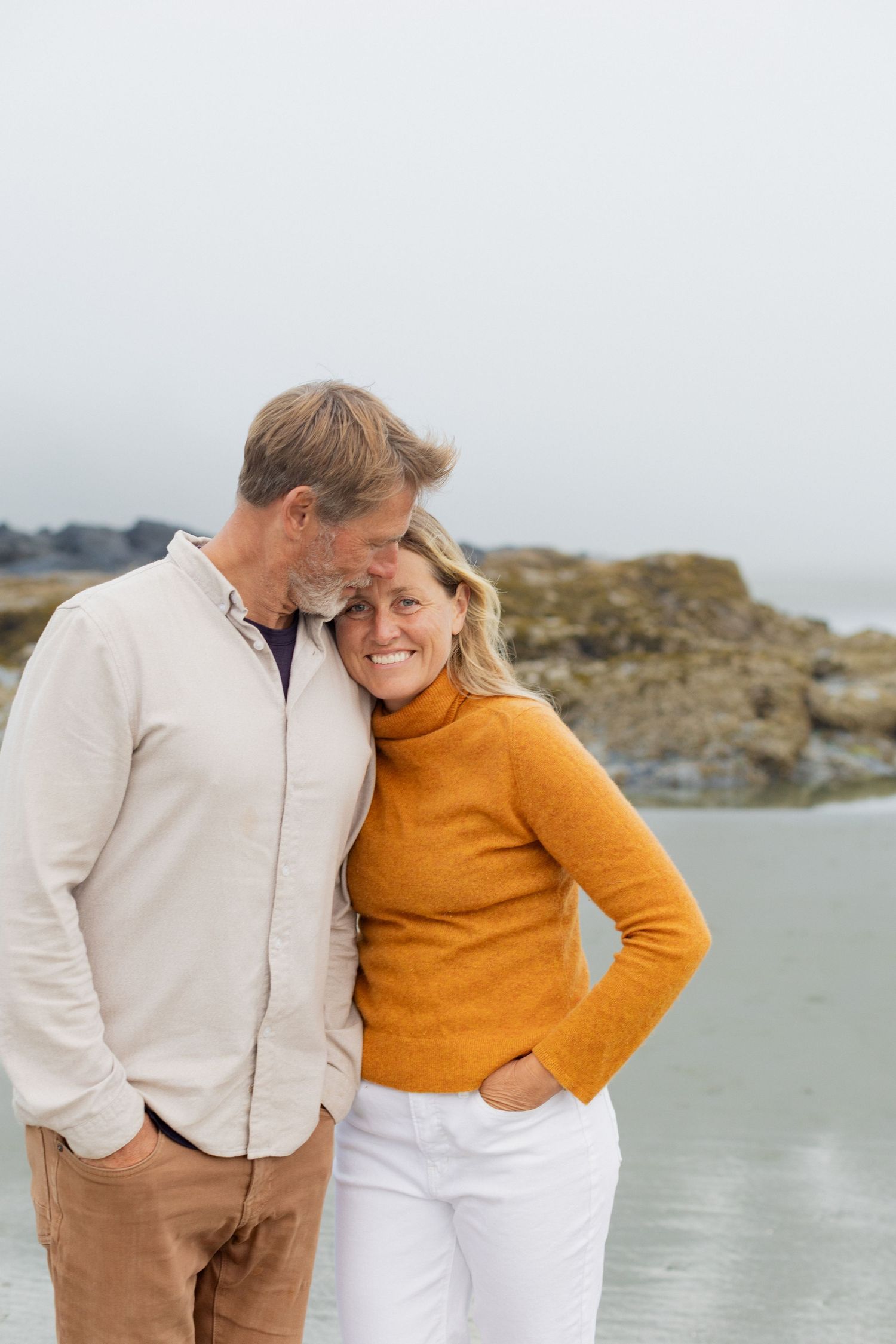 Man tenderly kissing smiling woman in orange sweater on rocky overcast coastal beach.