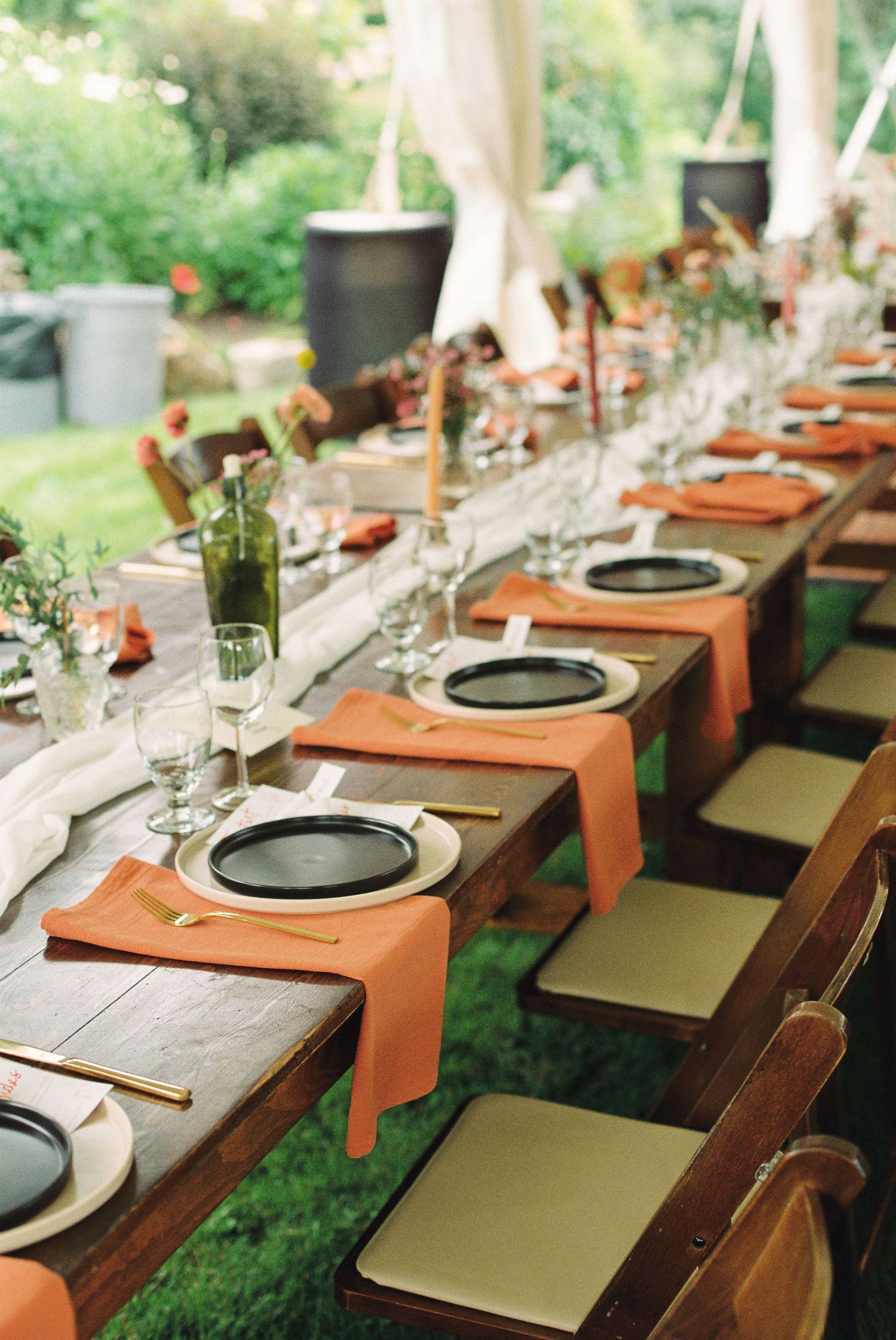 Elegant outdoor wedding reception table set with terracotta napkins, black plates, and gold cutlery.
