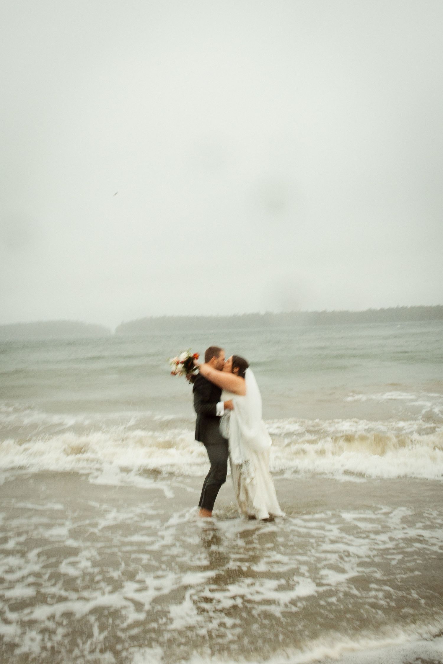 Bride and groom embrace and kiss while standing in ocean waves on a stormy wedding day.
