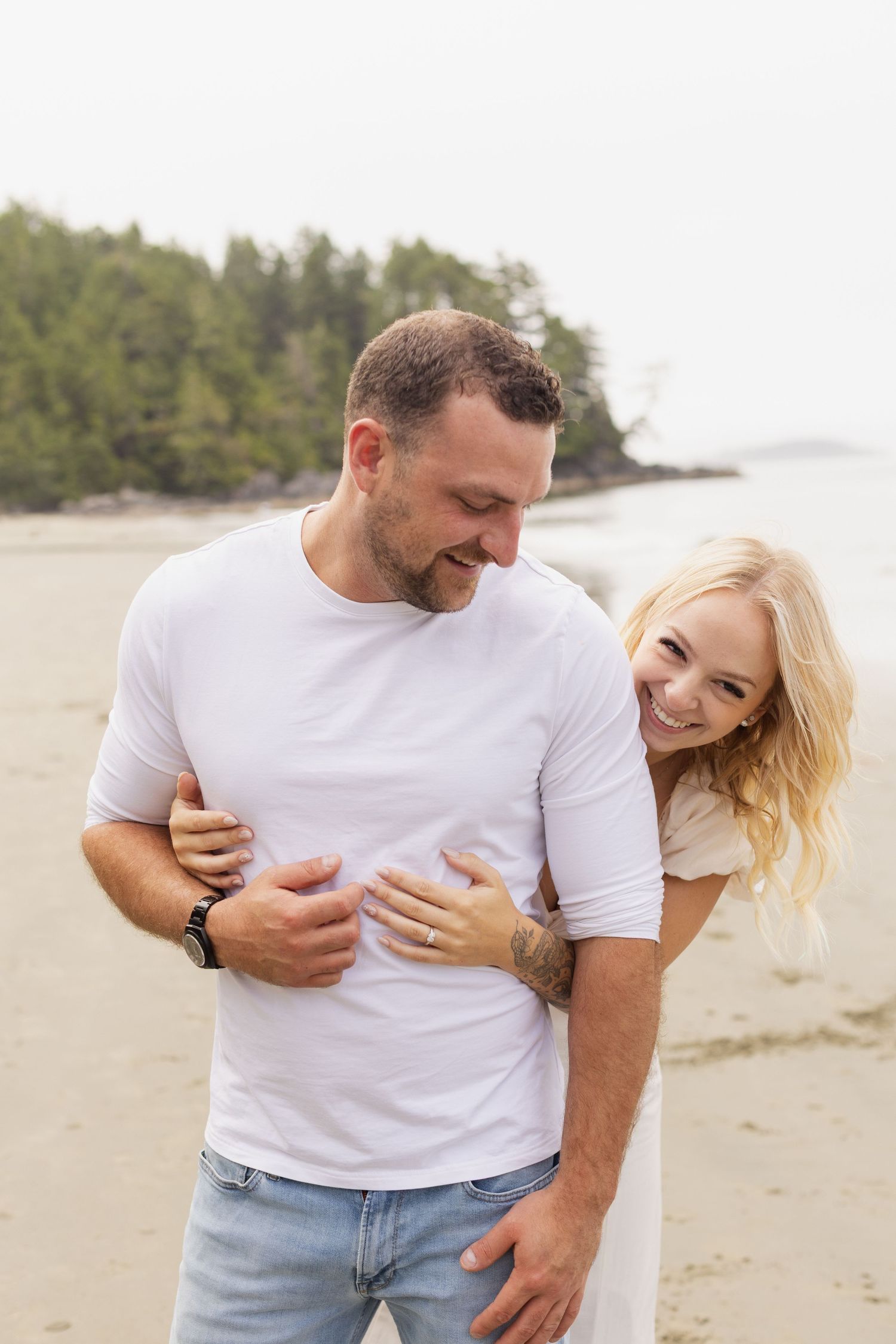 Woman in white dress playfully hugging man from behind on a sandy beach, both laughing.