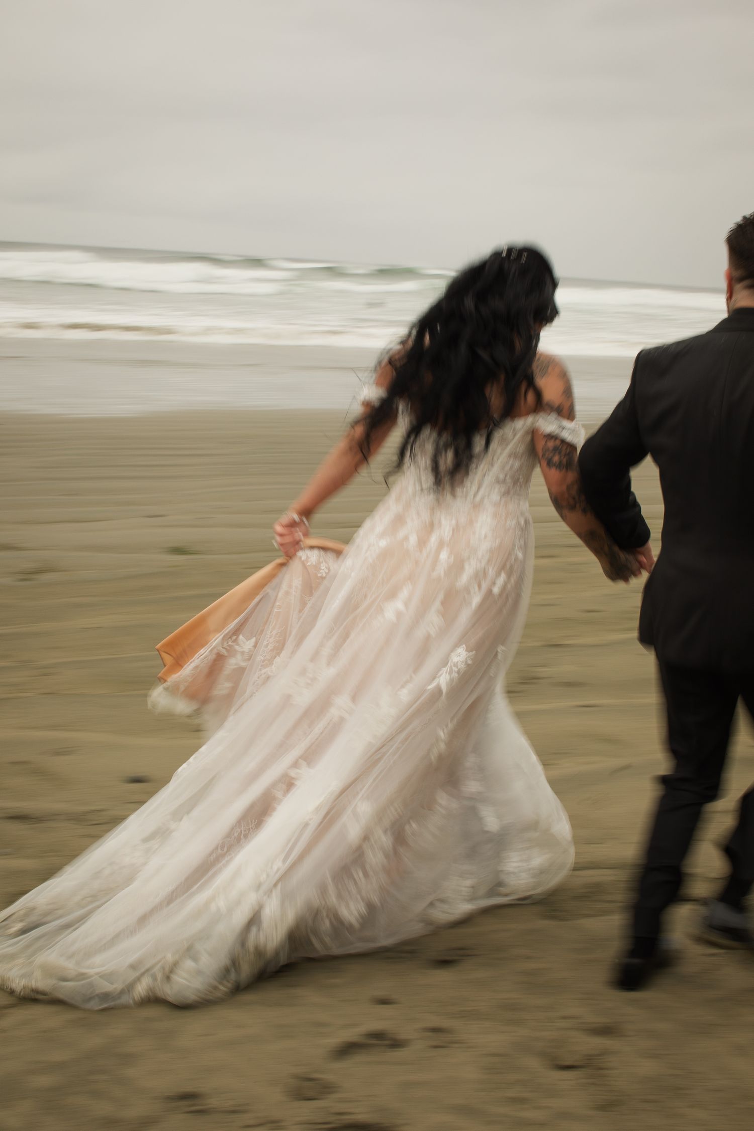 Bride in flowing lace gown running along a misty beach with groom in black suit on wedding day.