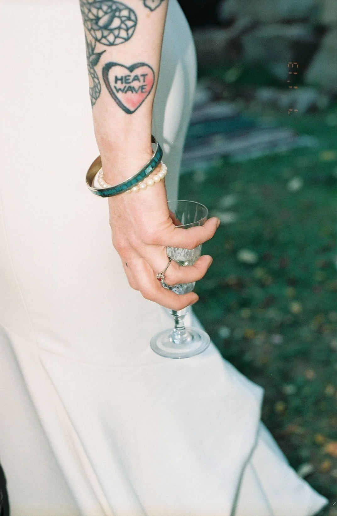 Close-up of tattooed bride holding small crystal glass, wearing gold and teal bangles with diamond ring.