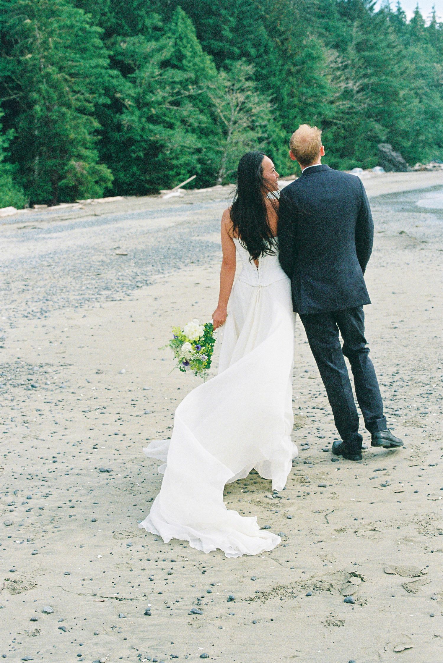 Bride in flowing white gown holding yellow bouquet walking hand in hand with groom on sandy beach.
