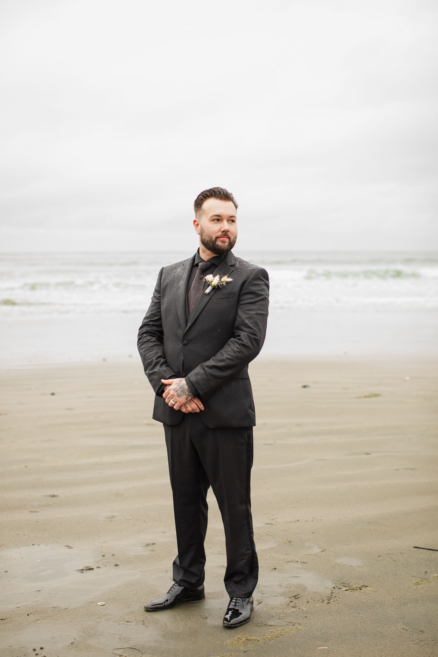 Groom in dark suit with floral boutonniere stands confidently on sandy beach with ocean backdrop.