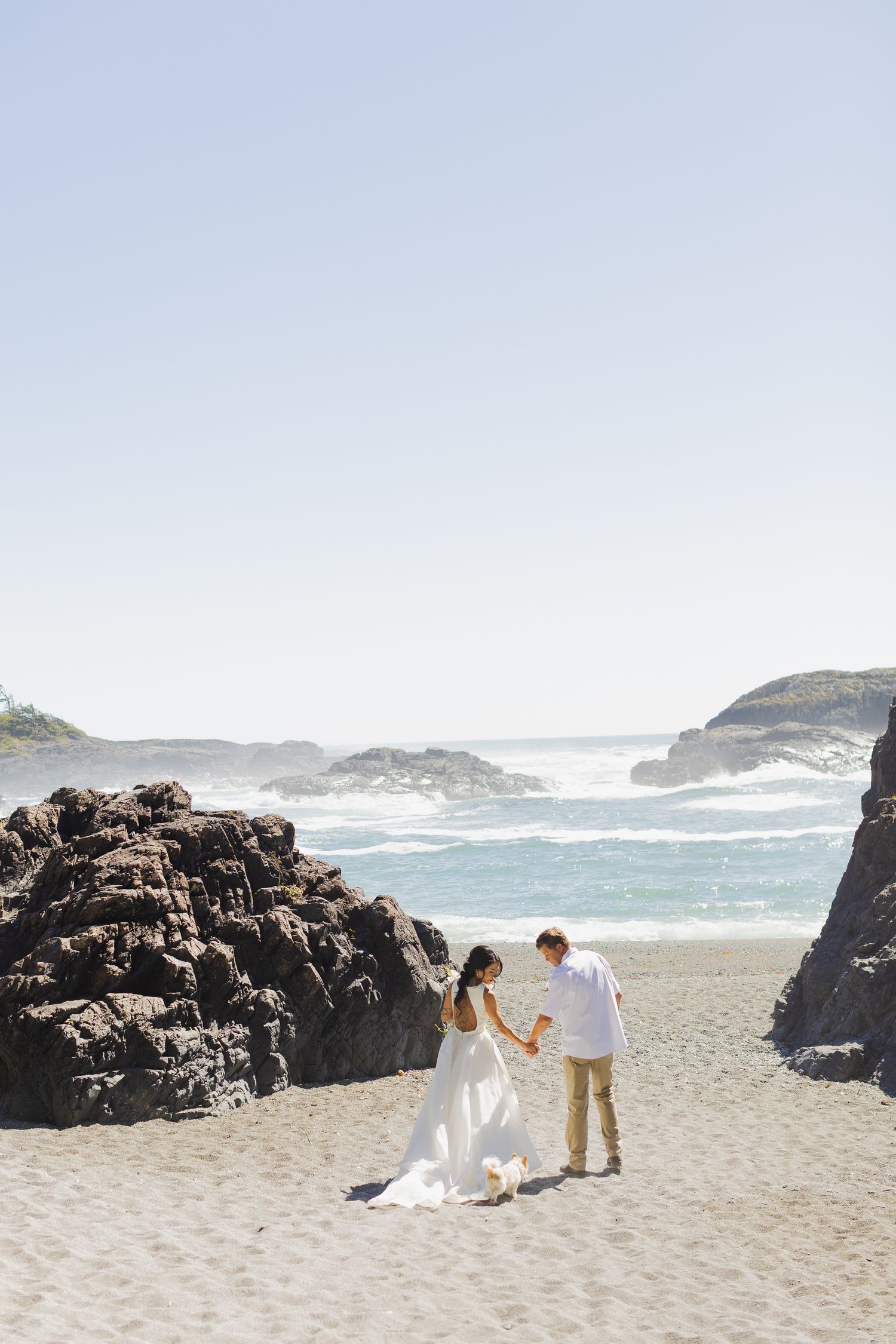 Bride and groom hold hands on a sunny sandy beach surrounded by dramatic rock formations and ocean waves.