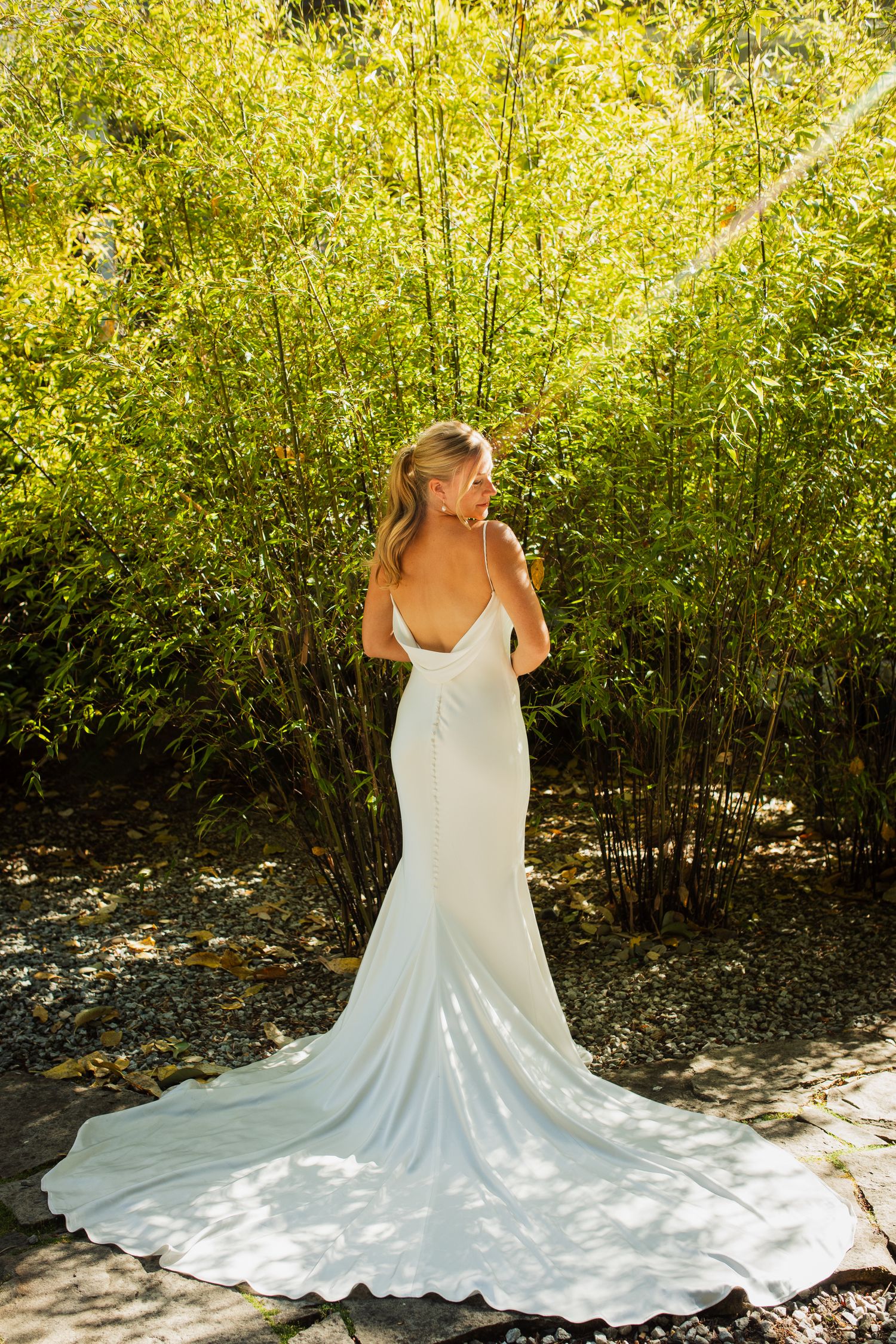 Bride in elegant backless white gown with train standing before lush green bamboo garden.
