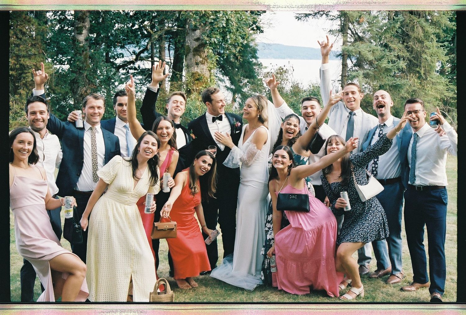 Joyful wedding party group photo outdoors with bride and groom surrounded by celebrating guests.