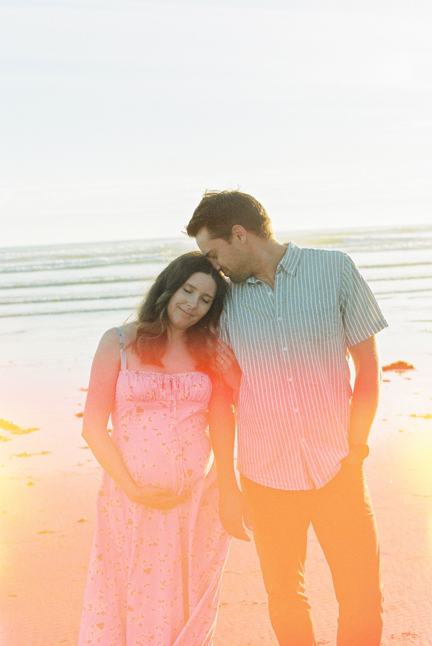 Pregnant couple standing close together on a sunlit beach, woman leaning on man's shoulder at golden hour.