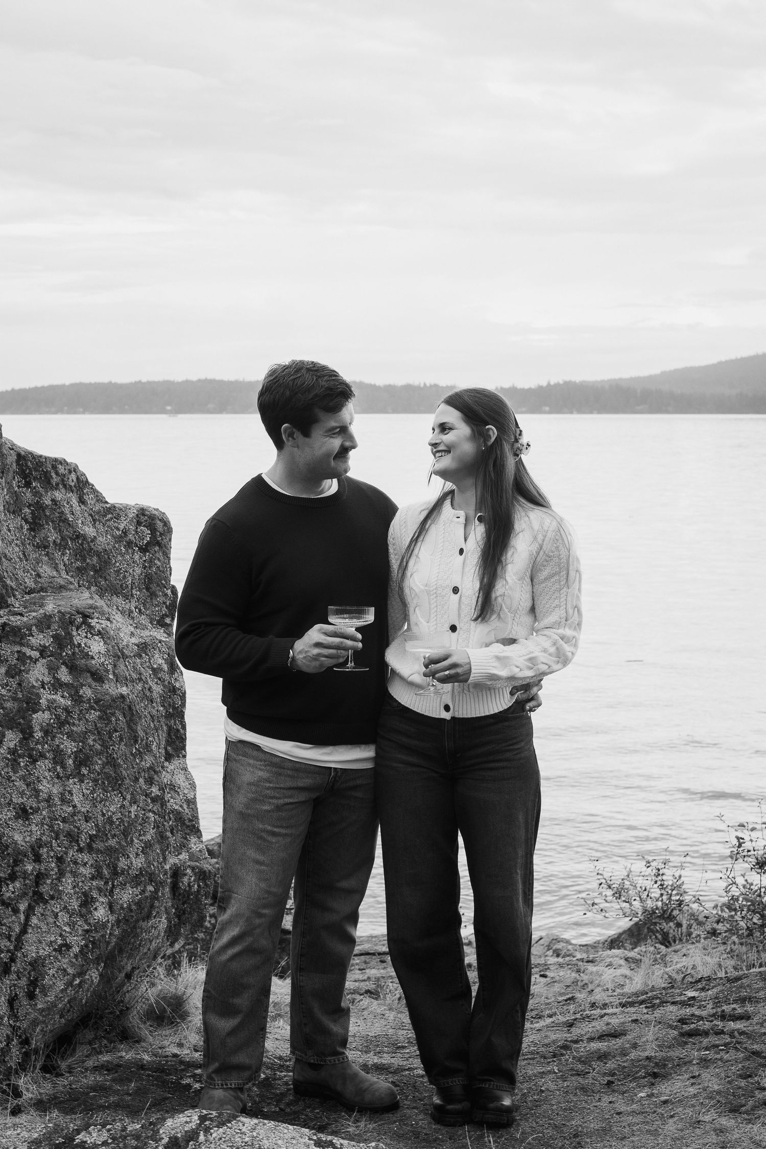 Black and white photo of a couple smiling at each other while holding wine glasses near a rocky waterfront.