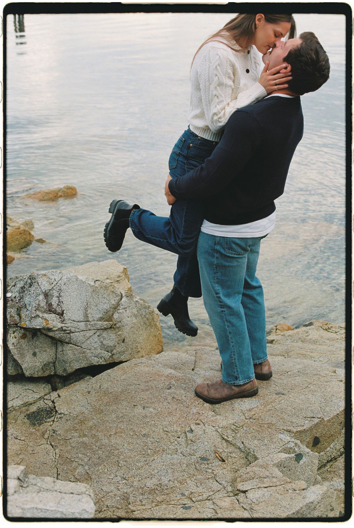 Man lifting woman for a kiss while standing on rocky shoreline beside a tranquil lake.
