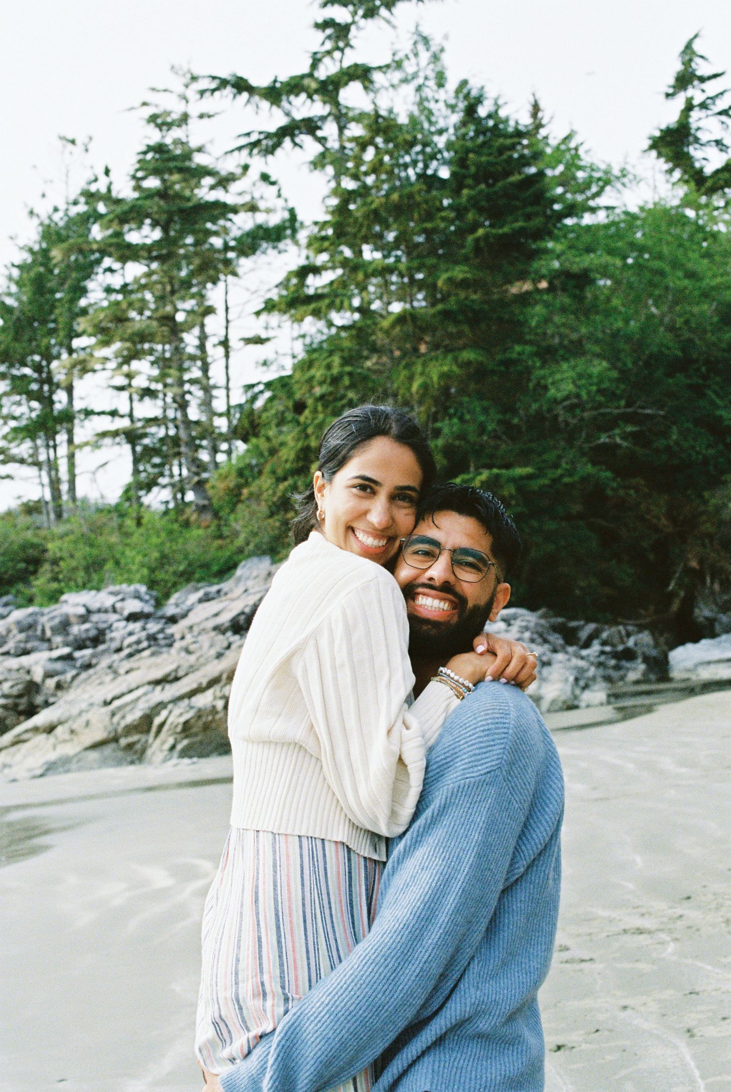 Smiling couple hugging on a sandy beach with rocky shoreline and pine trees.