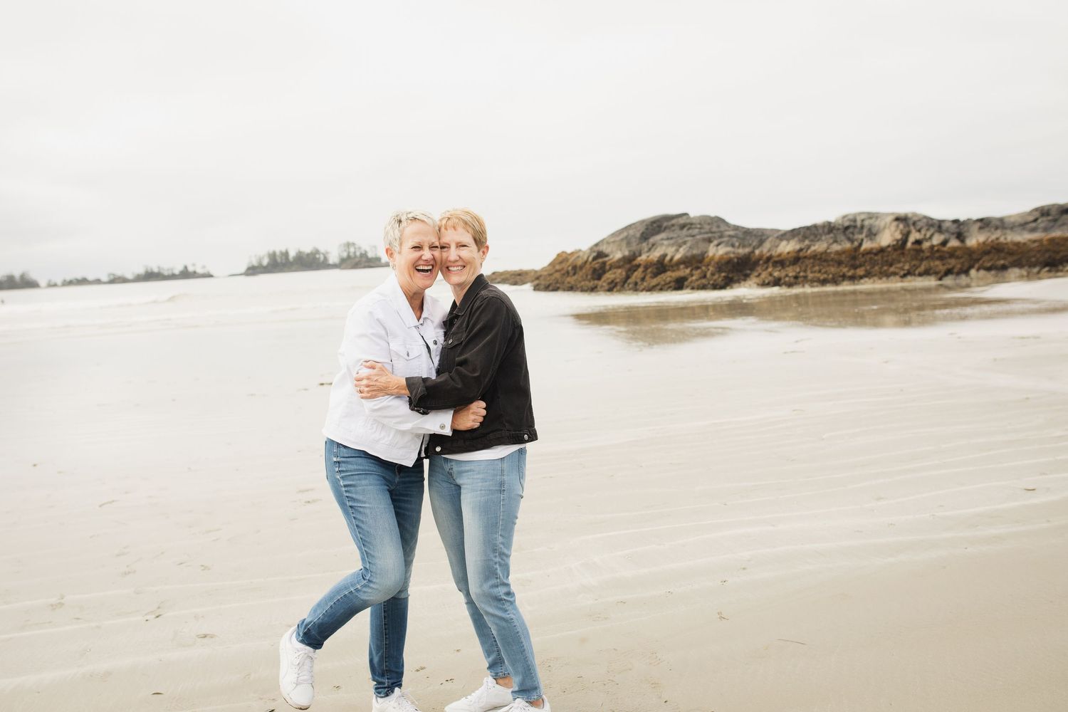 Two smiling mature women hugging and walking together on a sandy beach with rocky cliffs.