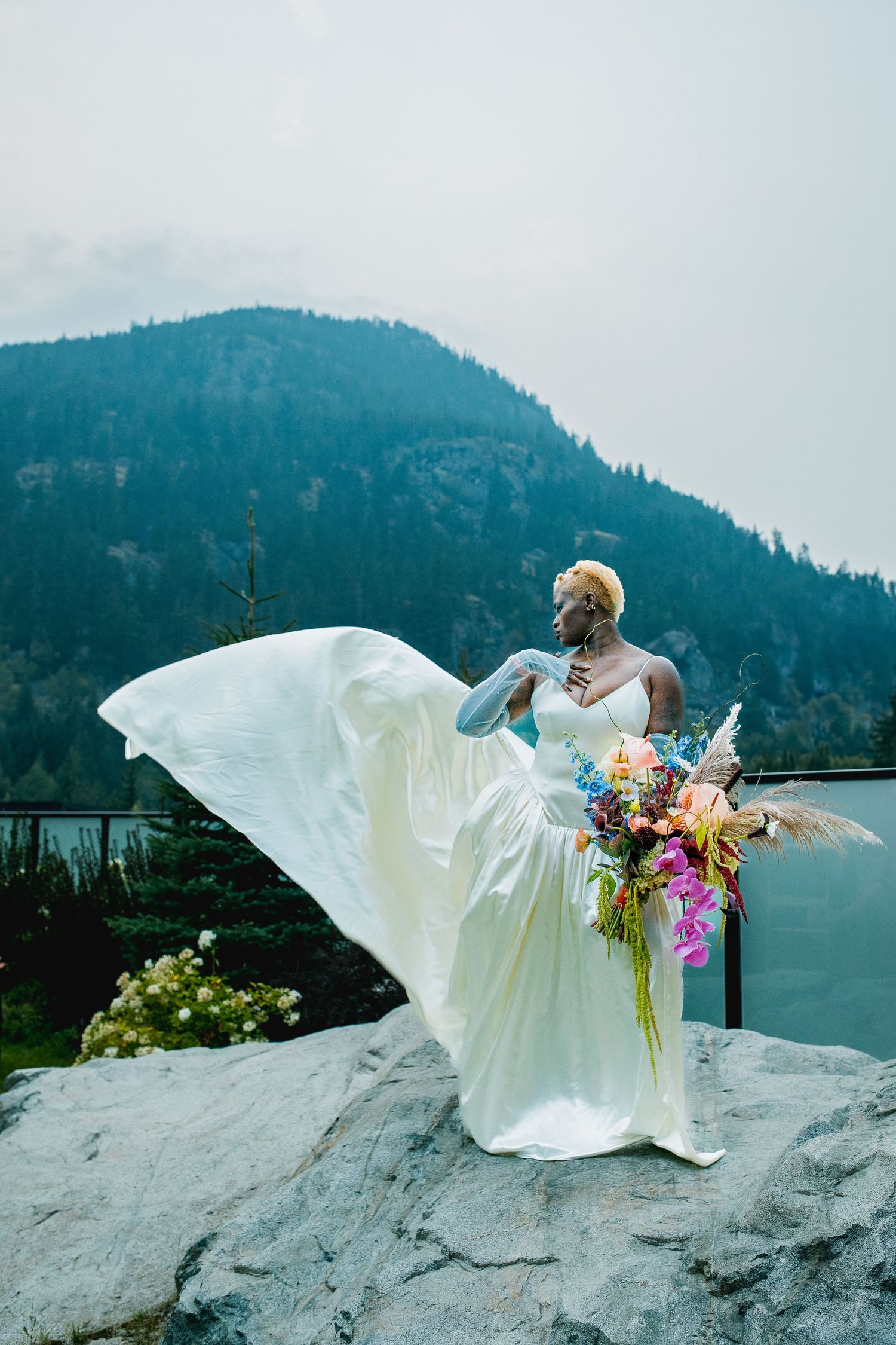 Black bride in flowing white wedding gown with colorful bouquet standing on rocks by mountain lake.