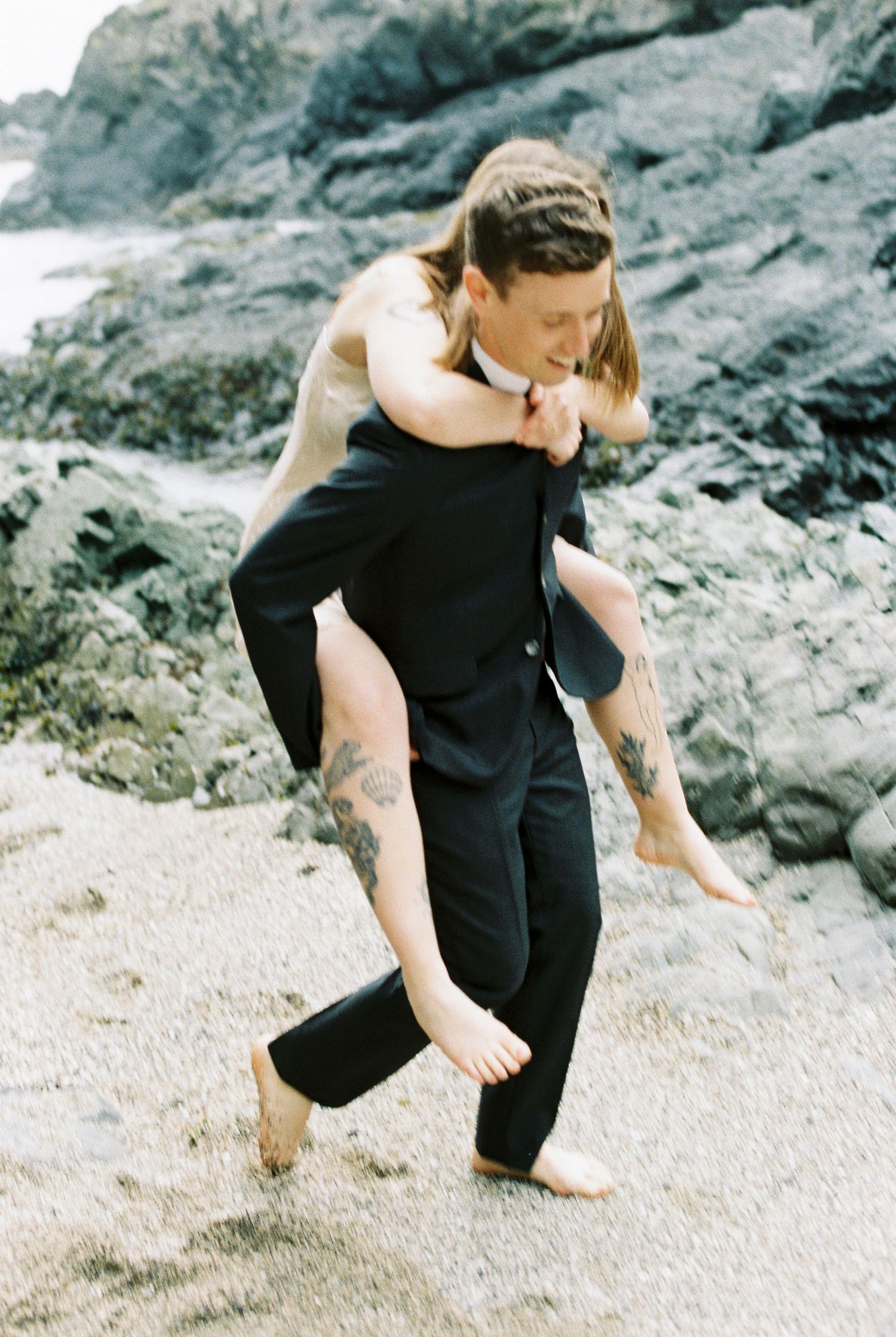 Groom carrying bride piggyback on a sandy beach with rocky cliffs in the background.