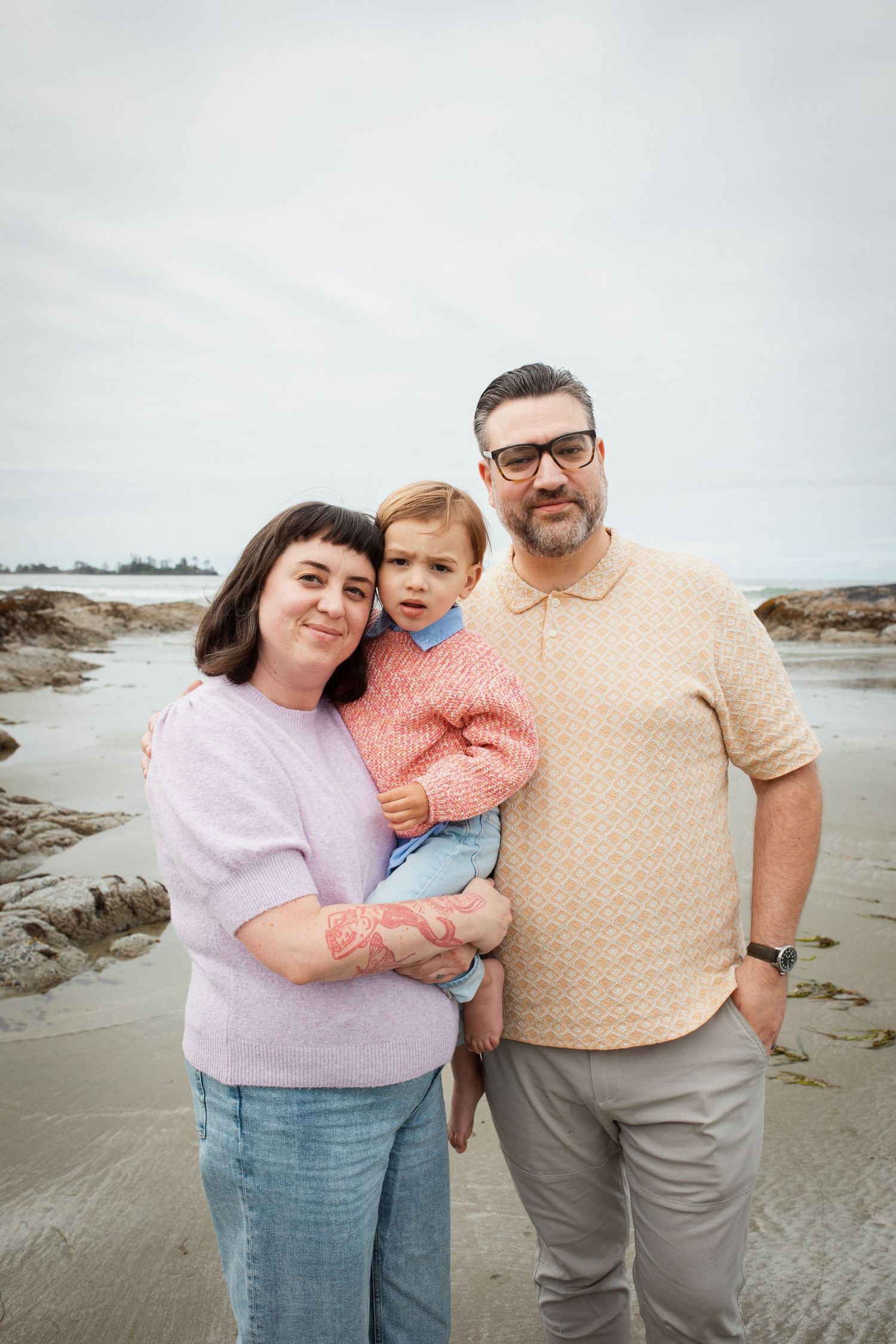 Family of three poses on a rocky beach, parents holding toddler, overcast sky backdrop.
