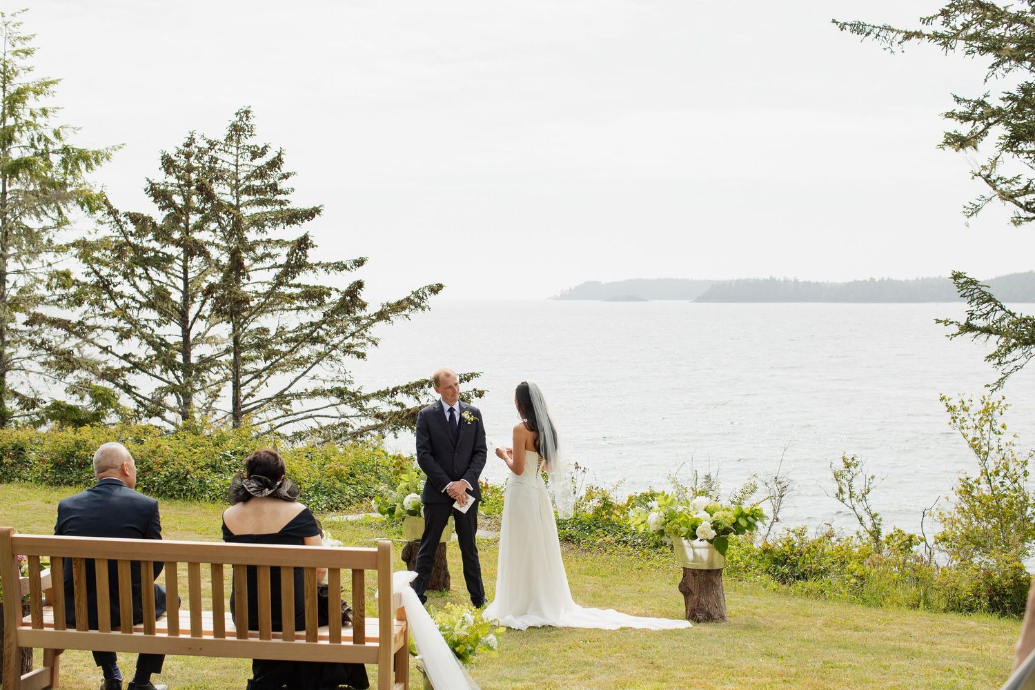 Intimate outdoor wedding ceremony with couple at altar overlooking ocean, guests seated on wooden bench.