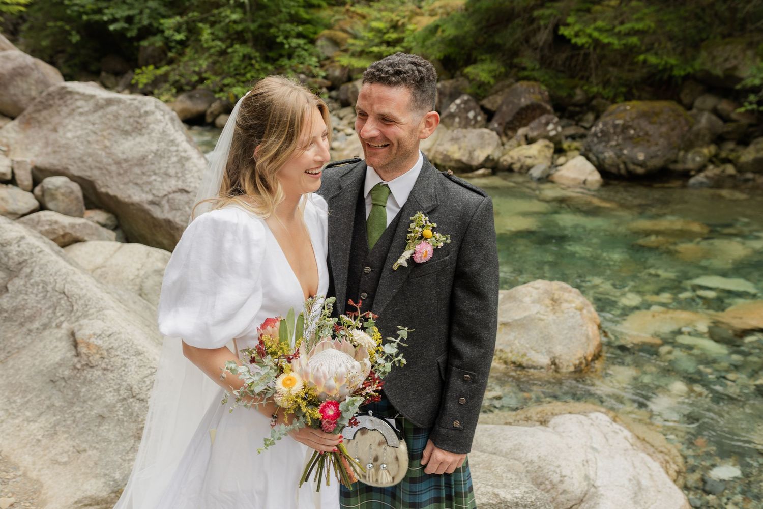 Smiling bride and groom posing by a rocky mountain stream, bride holding a colorful wildflower bouquet.
