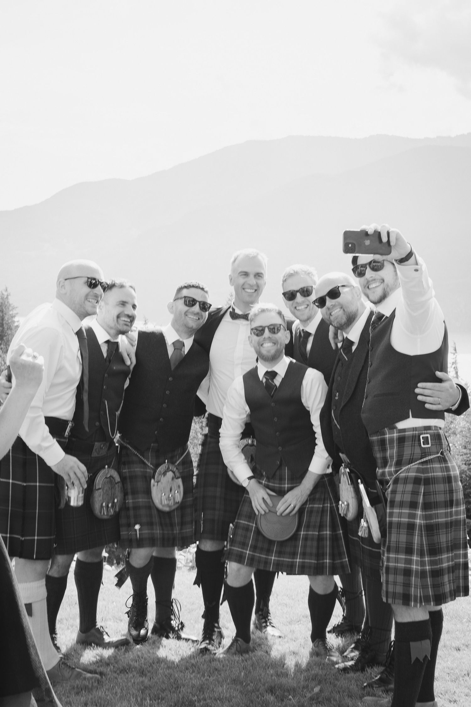 Group of groomsmen in matching Scottish kilts taking a fun selfie outdoors with mountains in background.