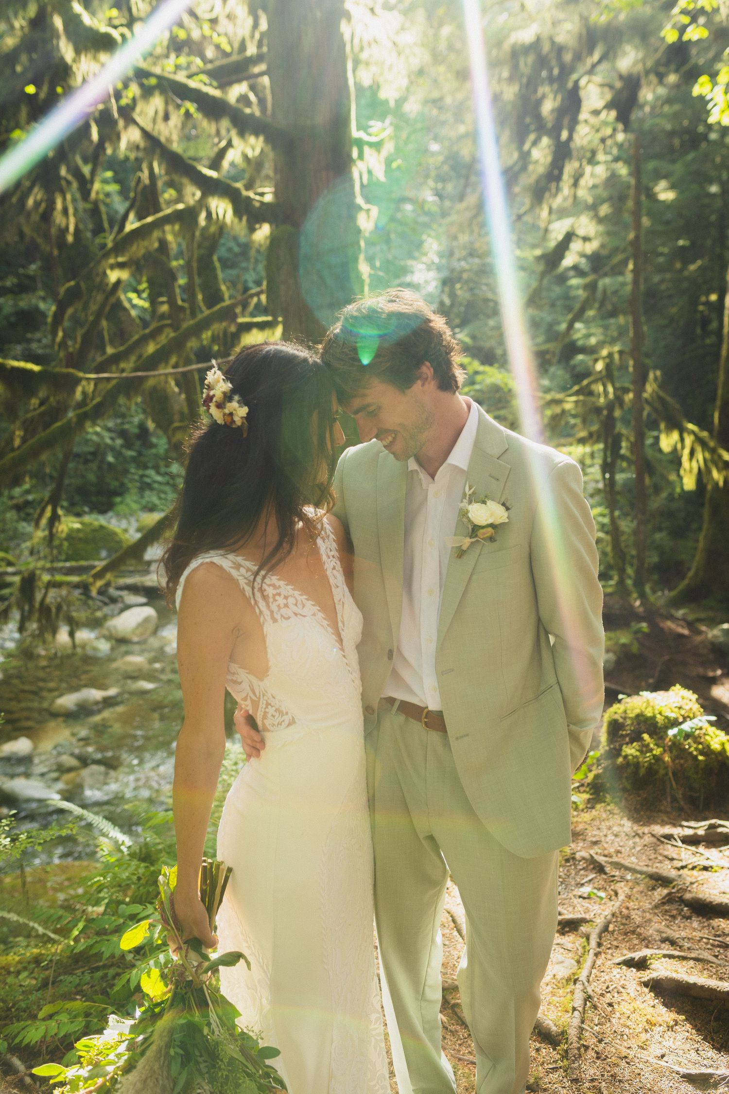 Bride and groom walk through sunlit forest with lens flare beside babbling creek.