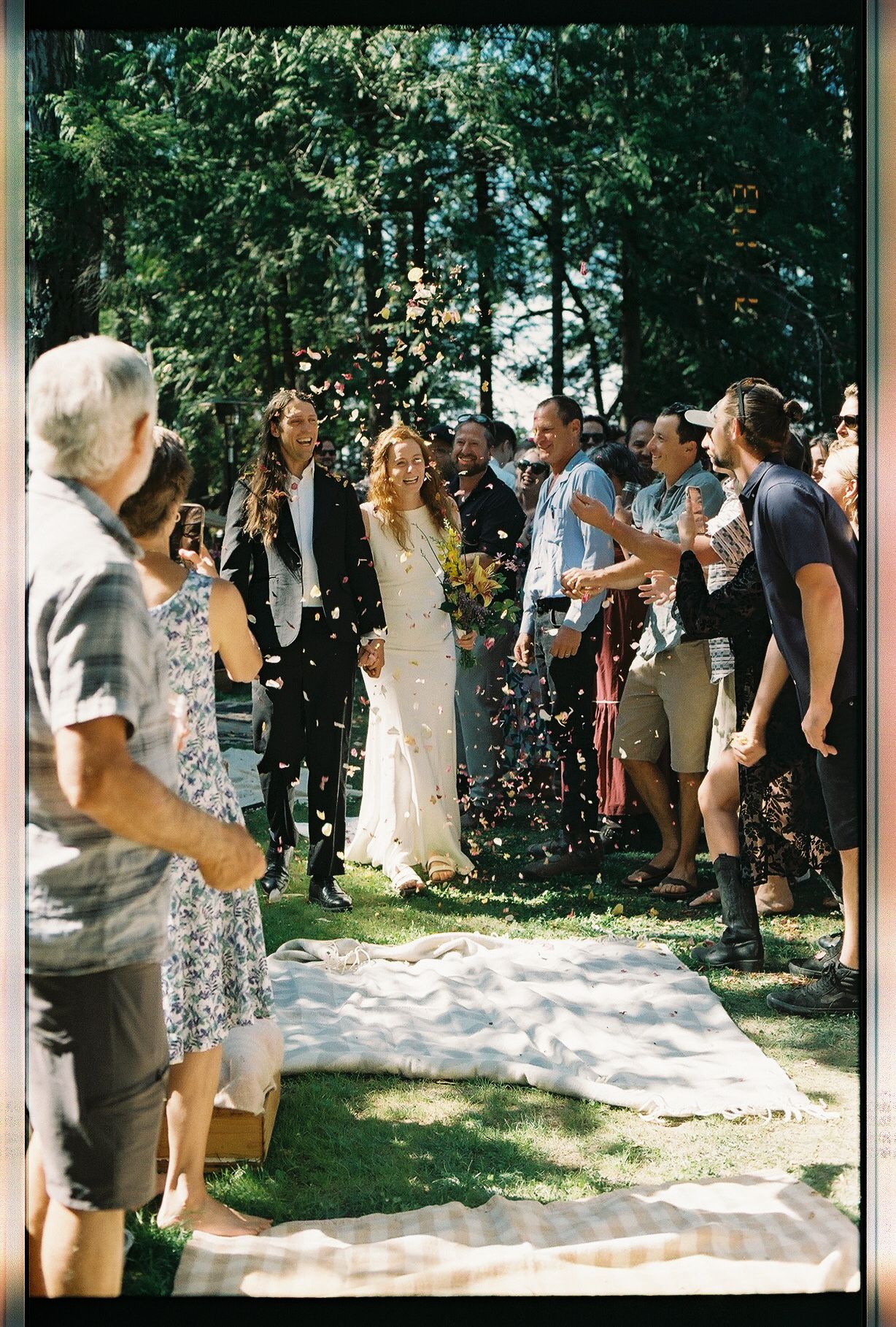 Bride and groom walking through confetti-throwing guests down outdoor aisle lined with trees.