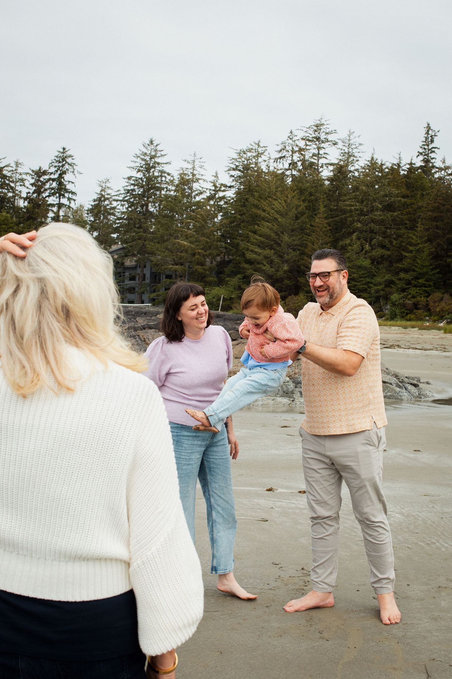 A happy family with a toddler laughs together on a scenic beach with tall evergreen trees behind.