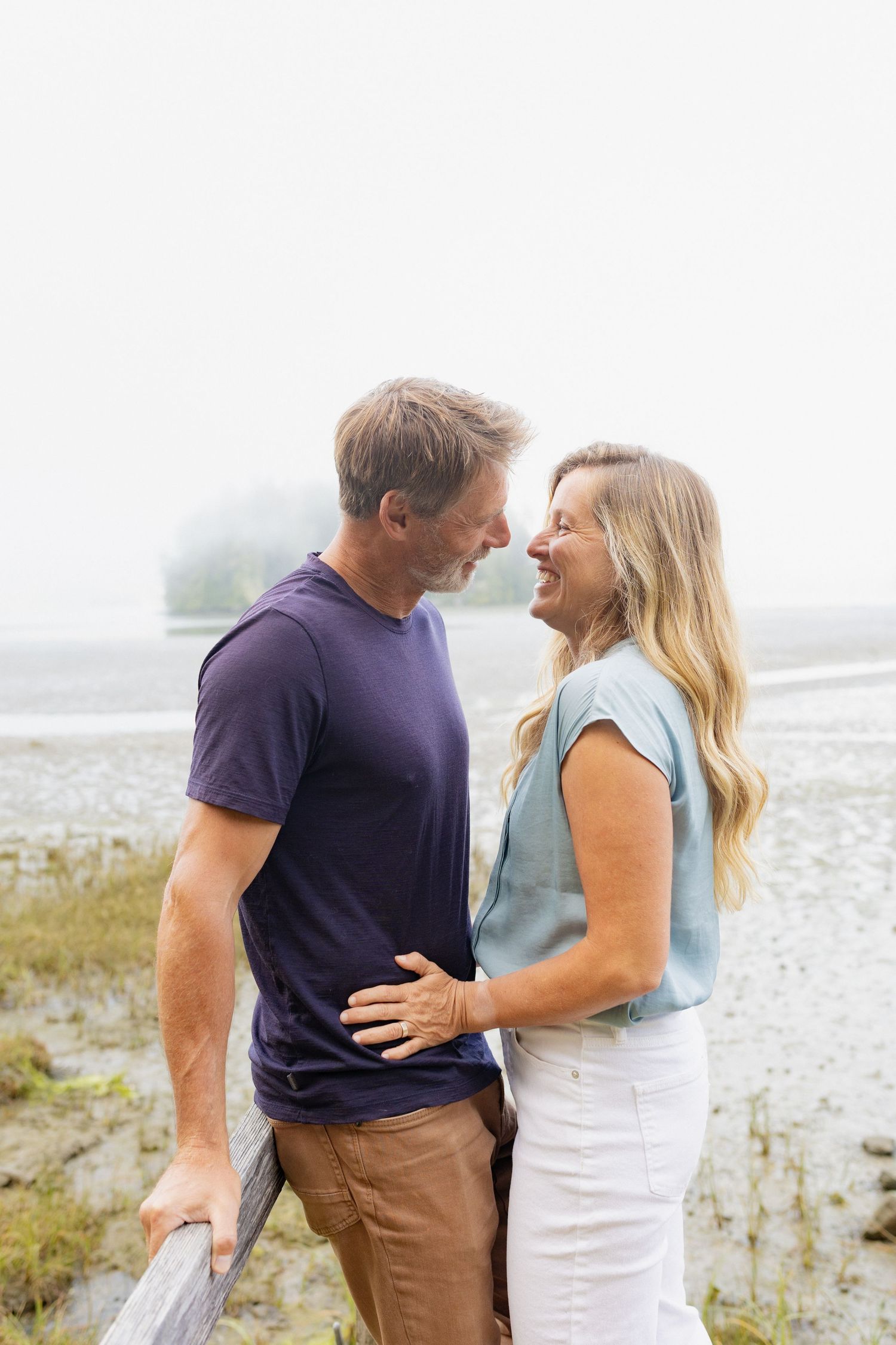 A middle-aged couple smiles at each other affectionately on a misty beach near tall grass.