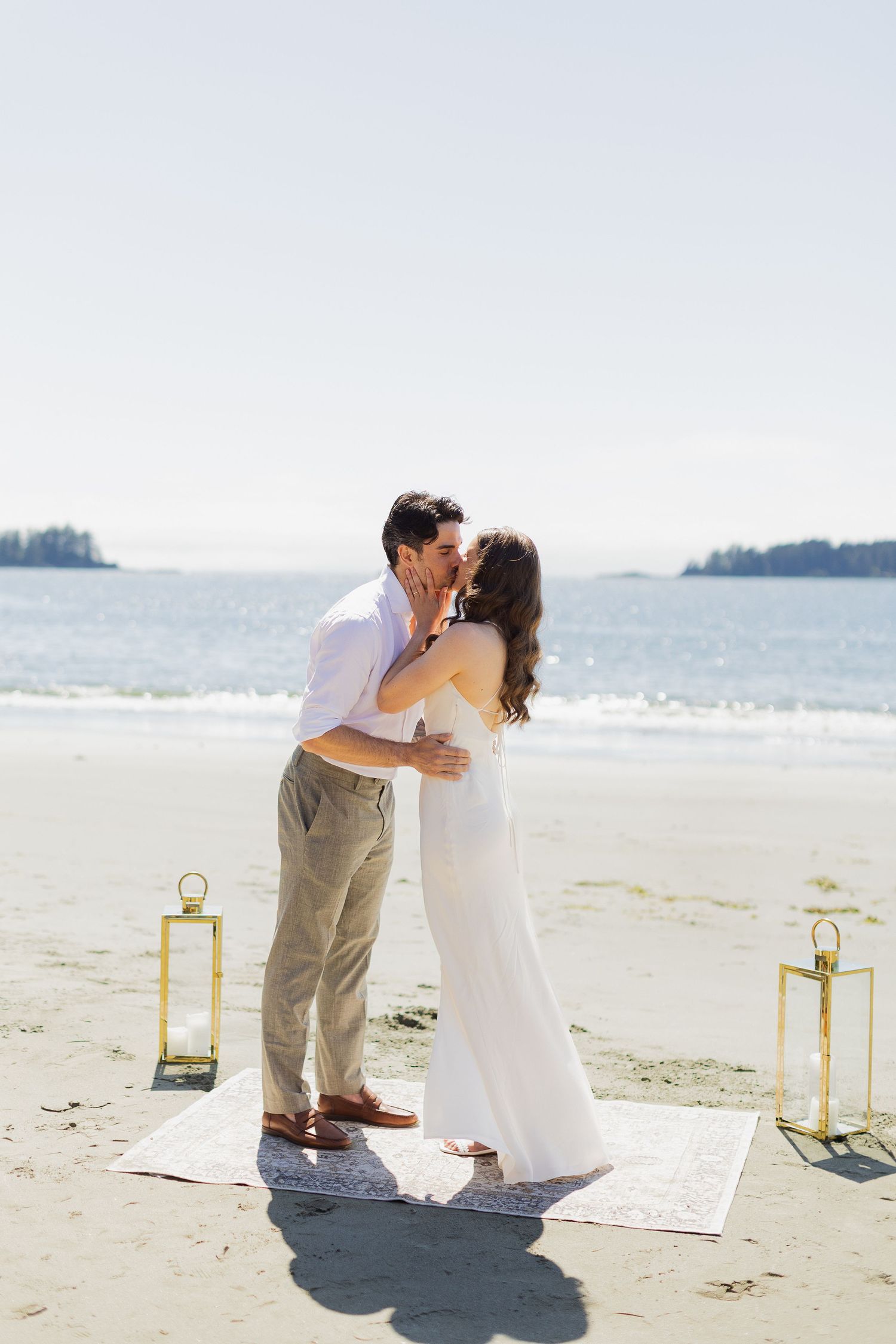 Bride and groom sharing first kiss on sandy beach with gold lanterns.