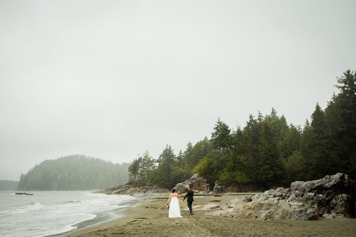 Bride and groom walk hand in hand along a misty beach surrounded by evergreen forest and rocky shores.