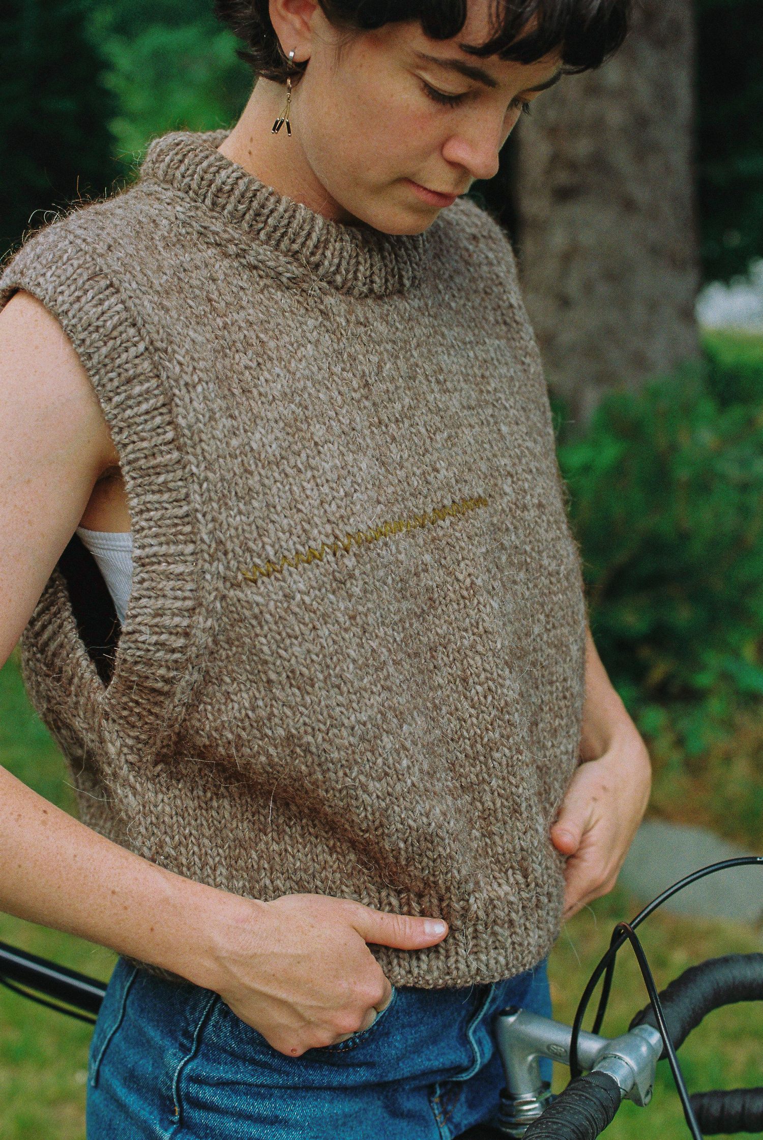 Woman in brown knit sleeveless vest and jeans stands near bicycle in outdoor setting.