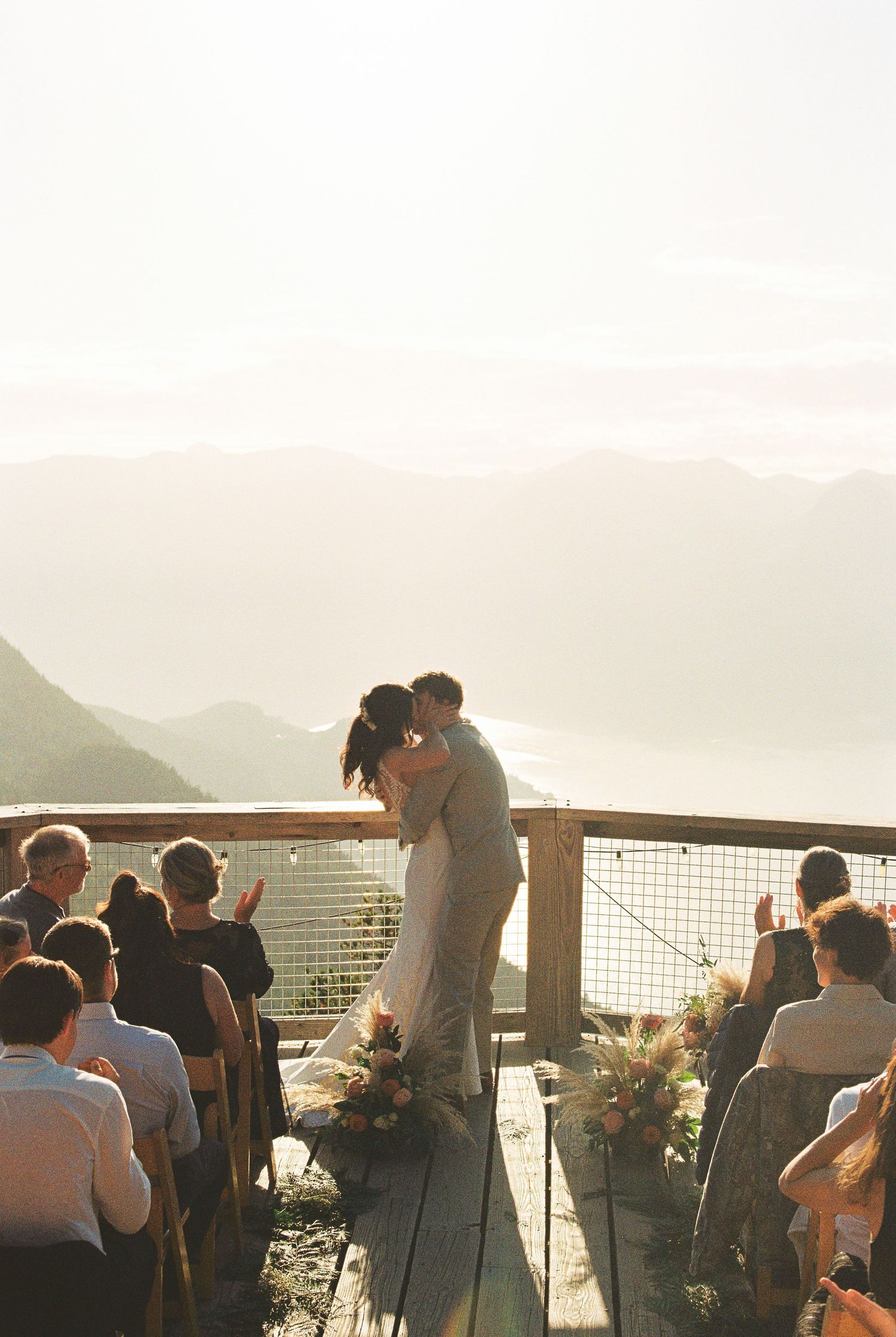 Couple shares first kiss at sunset mountain ceremony on wooden deck overlooking misty valley and peaks.