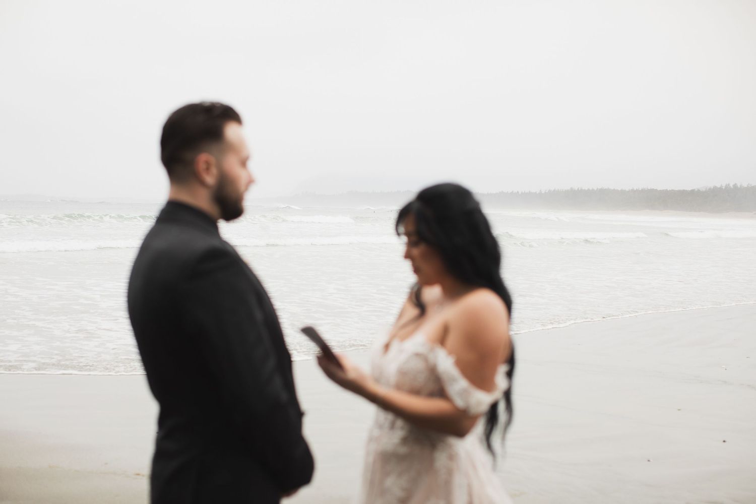 Bride reads wedding vows from phone while groom listens during intimate beach elopement ceremony.
