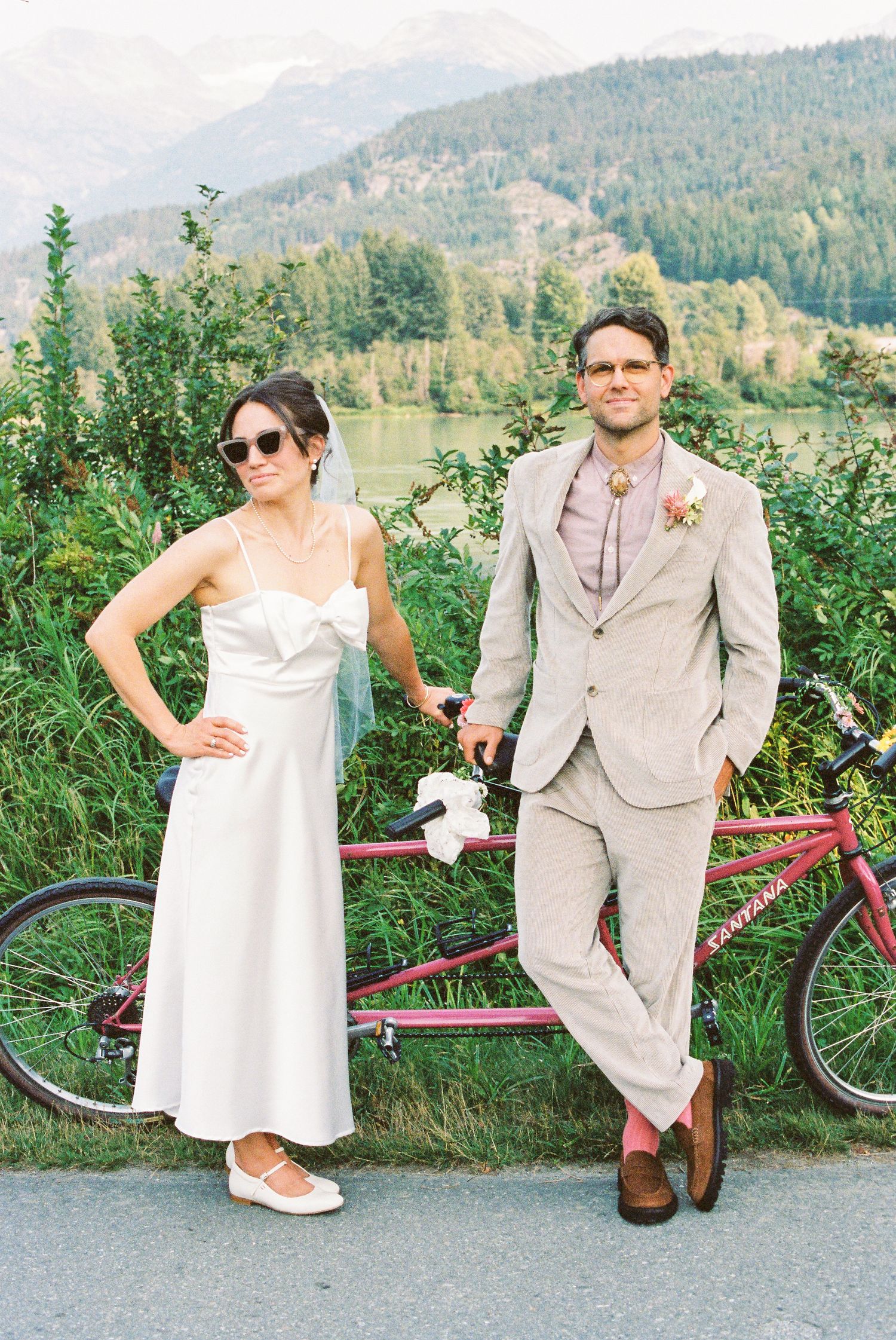 Bride and groom pose coolly beside a tandem bicycle with mountain and lake backdrop on their wedding day.