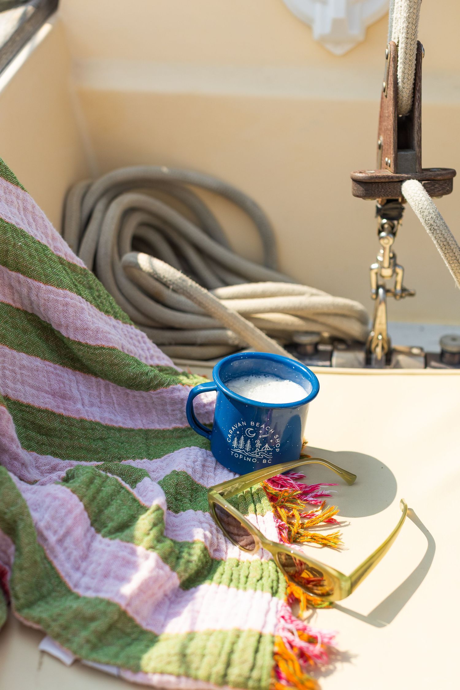 Blue enamel camp mug with frothy drink on striped blanket aboard sunny sailboat deck.