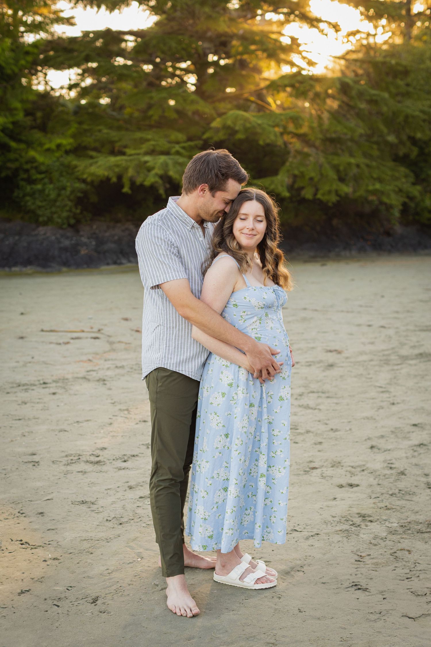 Expectant couple embracing on a beach at sunset, man tenderly holding pregnant woman's baby bump.