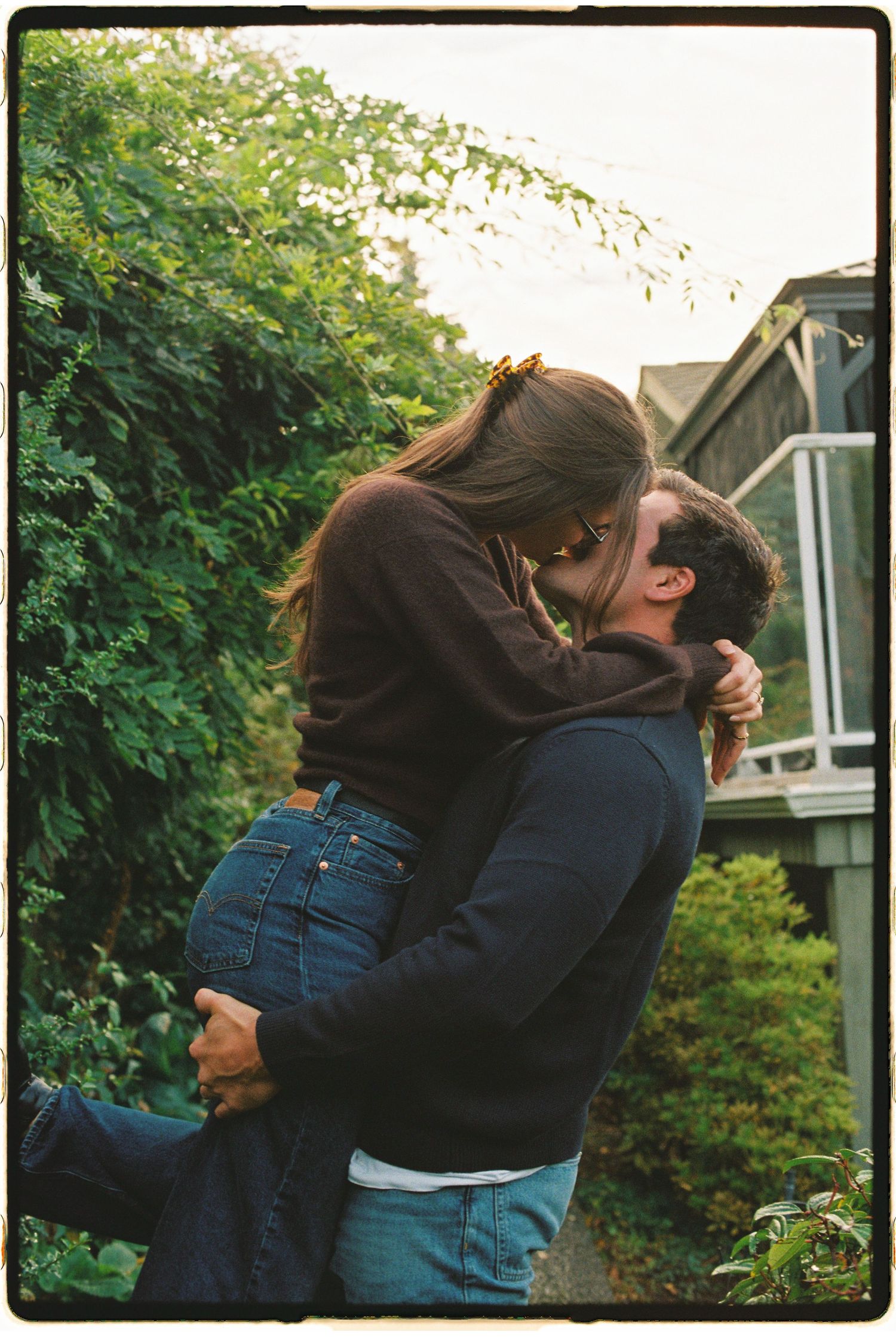 Man lifting woman for a romantic kiss in front of a green garden and house exterior.
