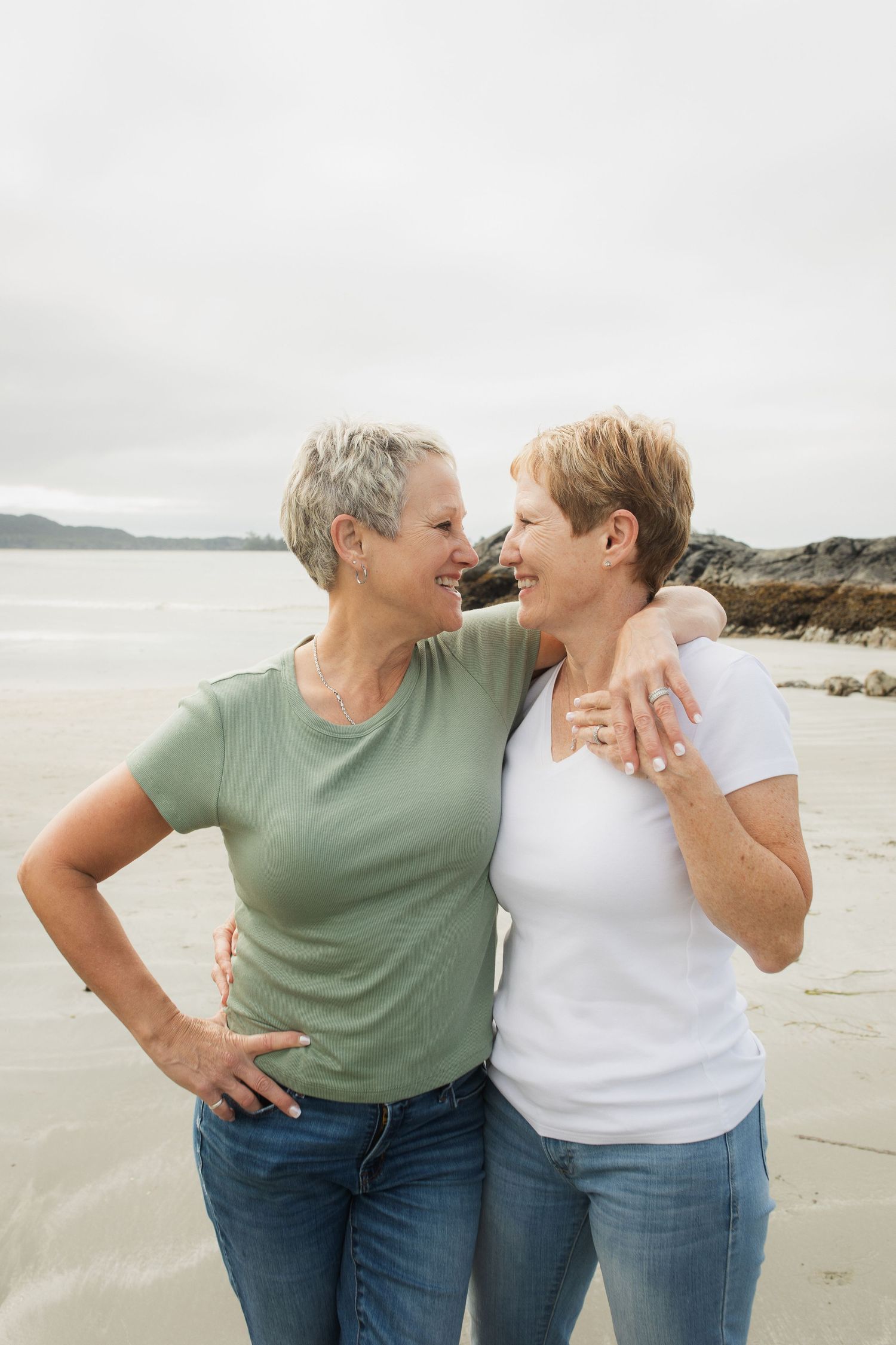 Two mature women with short hair embrace and smile at each other on a sandy beach on an overcast day.