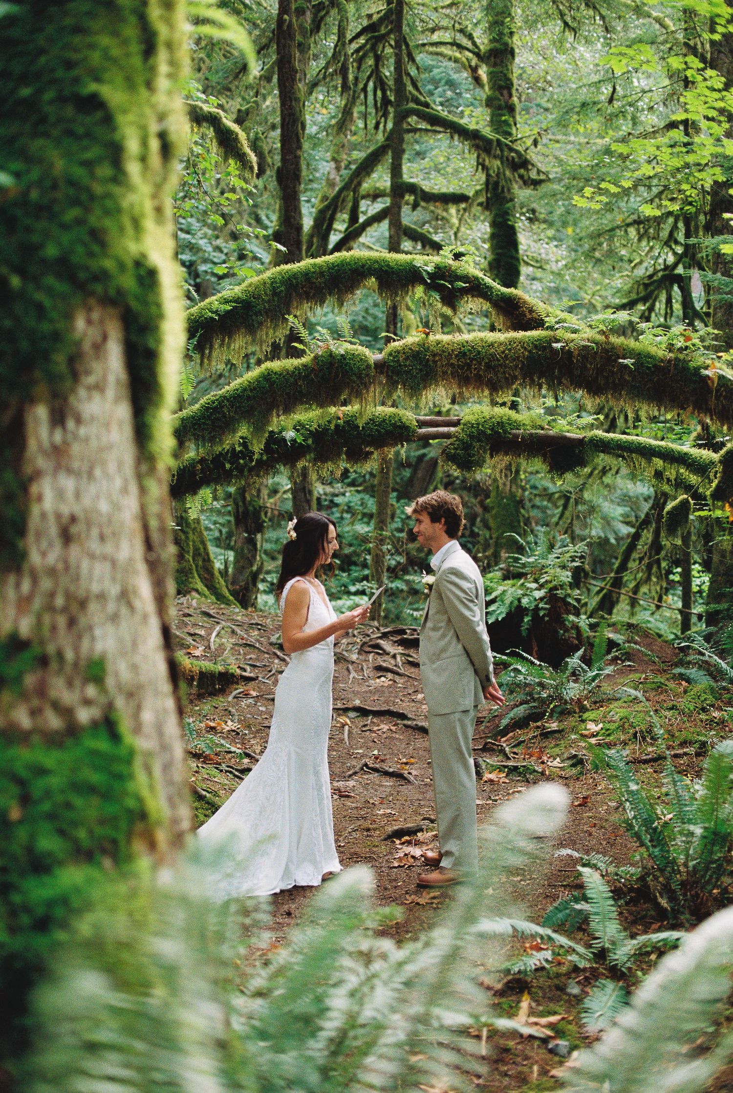 Bride and groom facing each other on forest path surrounded by moss-covered trees and ferns.