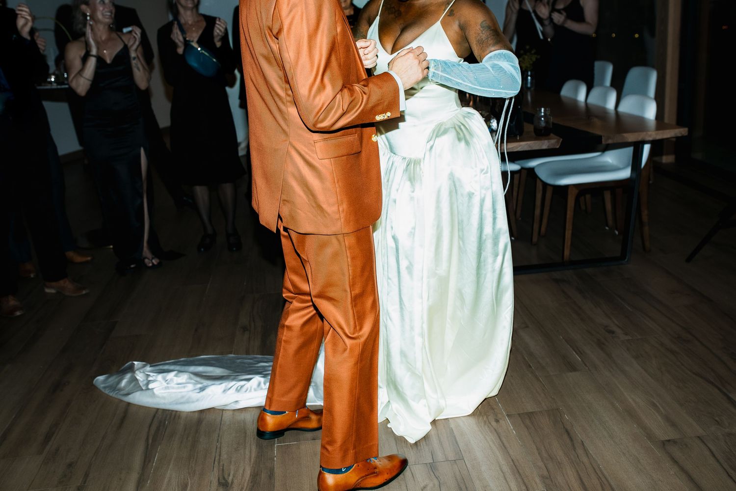 Couple sharing first dance, groom in orange suit and bride in white gown on wooden dance floor.