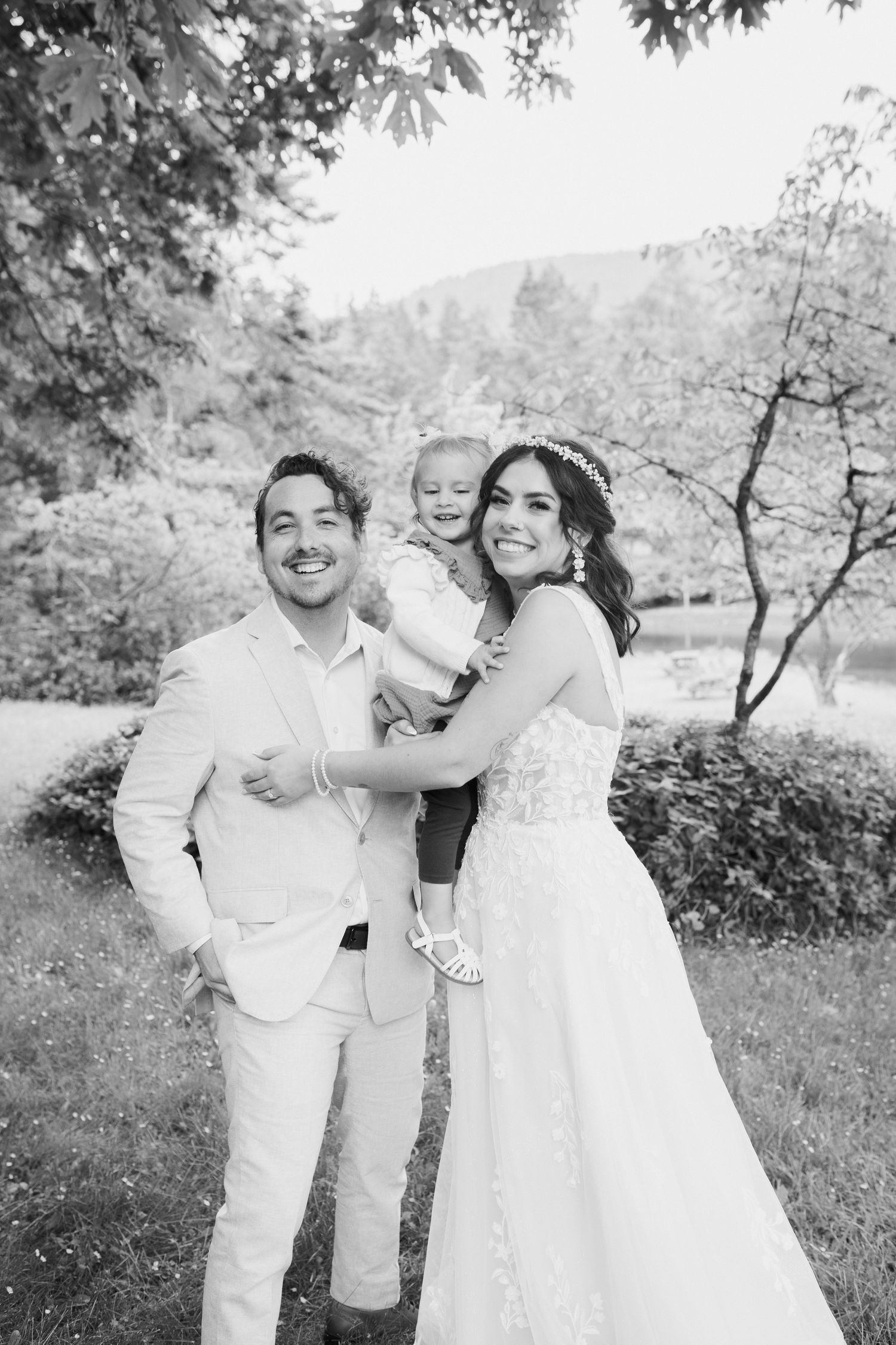 Black and white photo of smiling bride and groom holding young child at outdoor wedding.