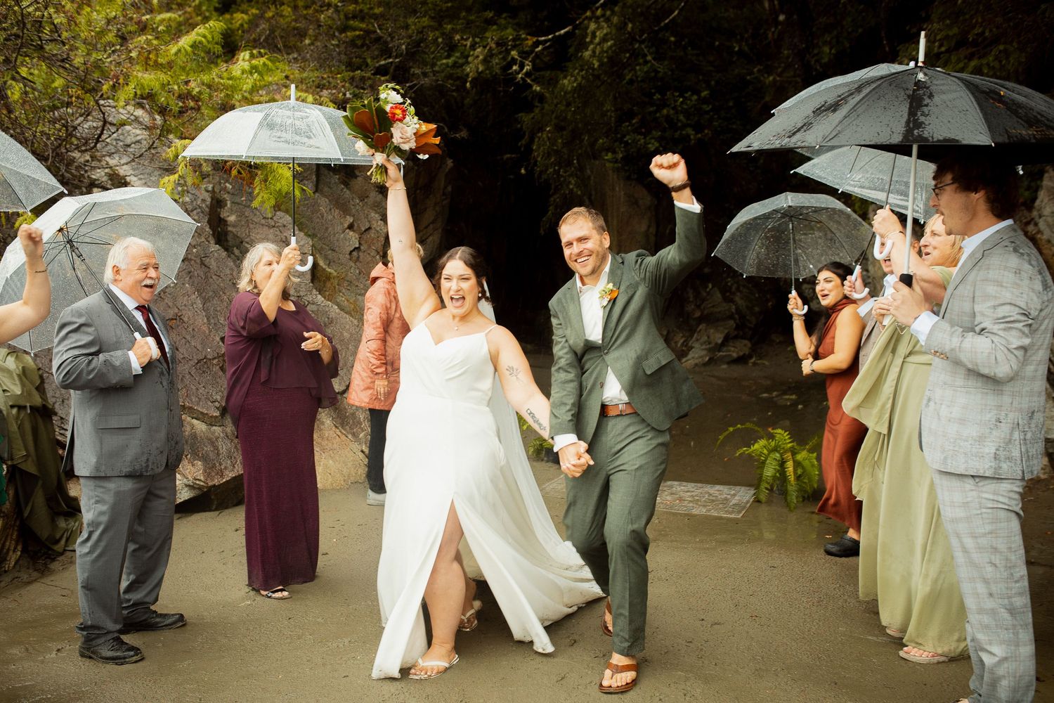 Joyful newlyweds celebrate recessional with raised arms surrounded by guests holding clear umbrellas.