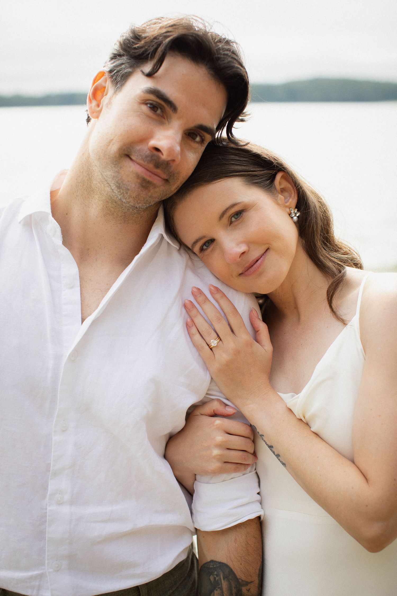 Couple in wedding attire pose closely together with serene lake and hills in background.