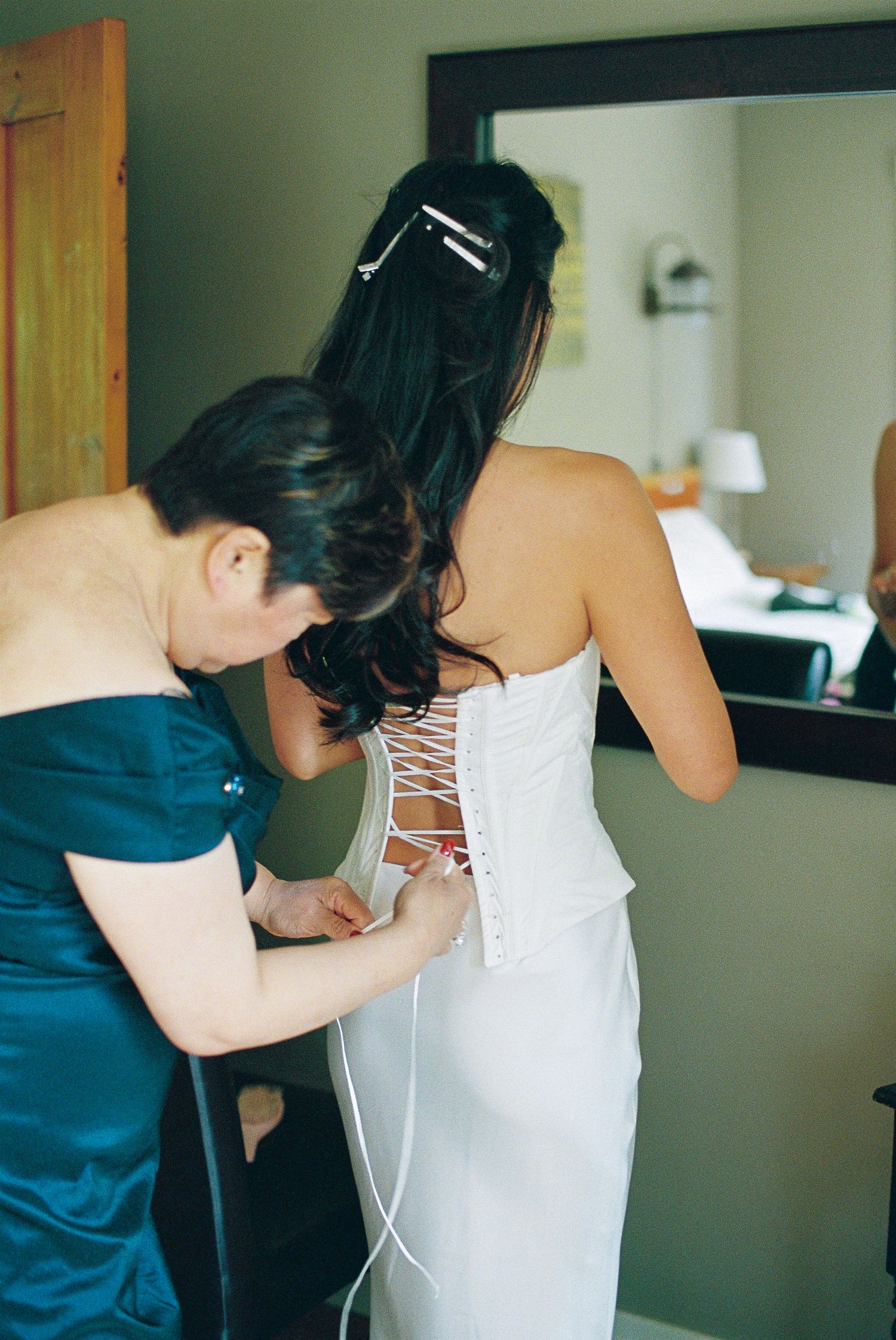 Bridesmaid in teal dress laces up the corset back of bride's white strapless wedding gown indoors.