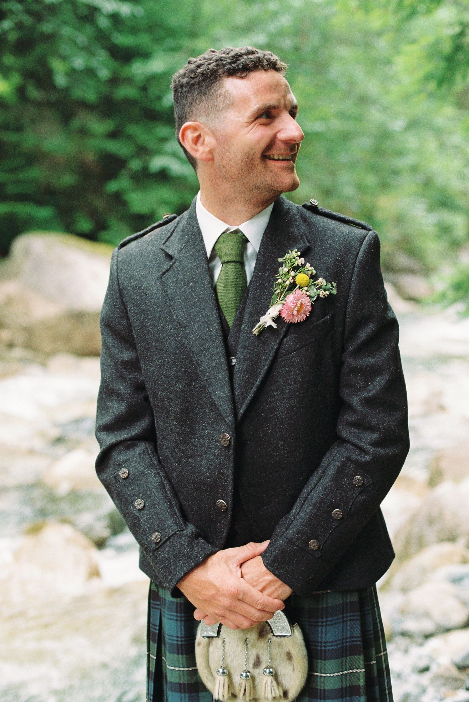 Smiling groom in Scottish tweed jacket and tartan kilt with floral boutonniere outdoors.