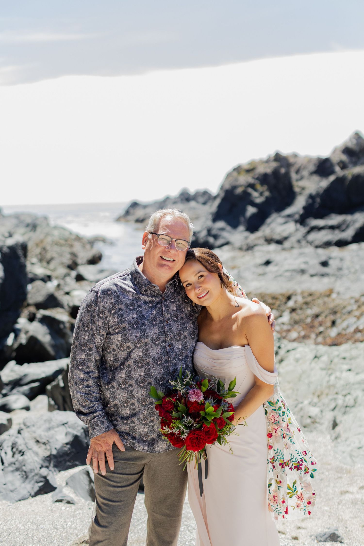 Smiling bride holding red bouquet poses with older man against rocky coastal backdrop.