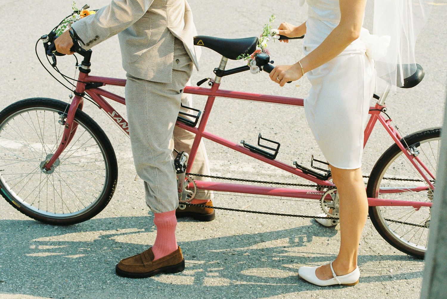 Bride and groom standing beside a red tandem bicycle on a sunny street on their wedding day.