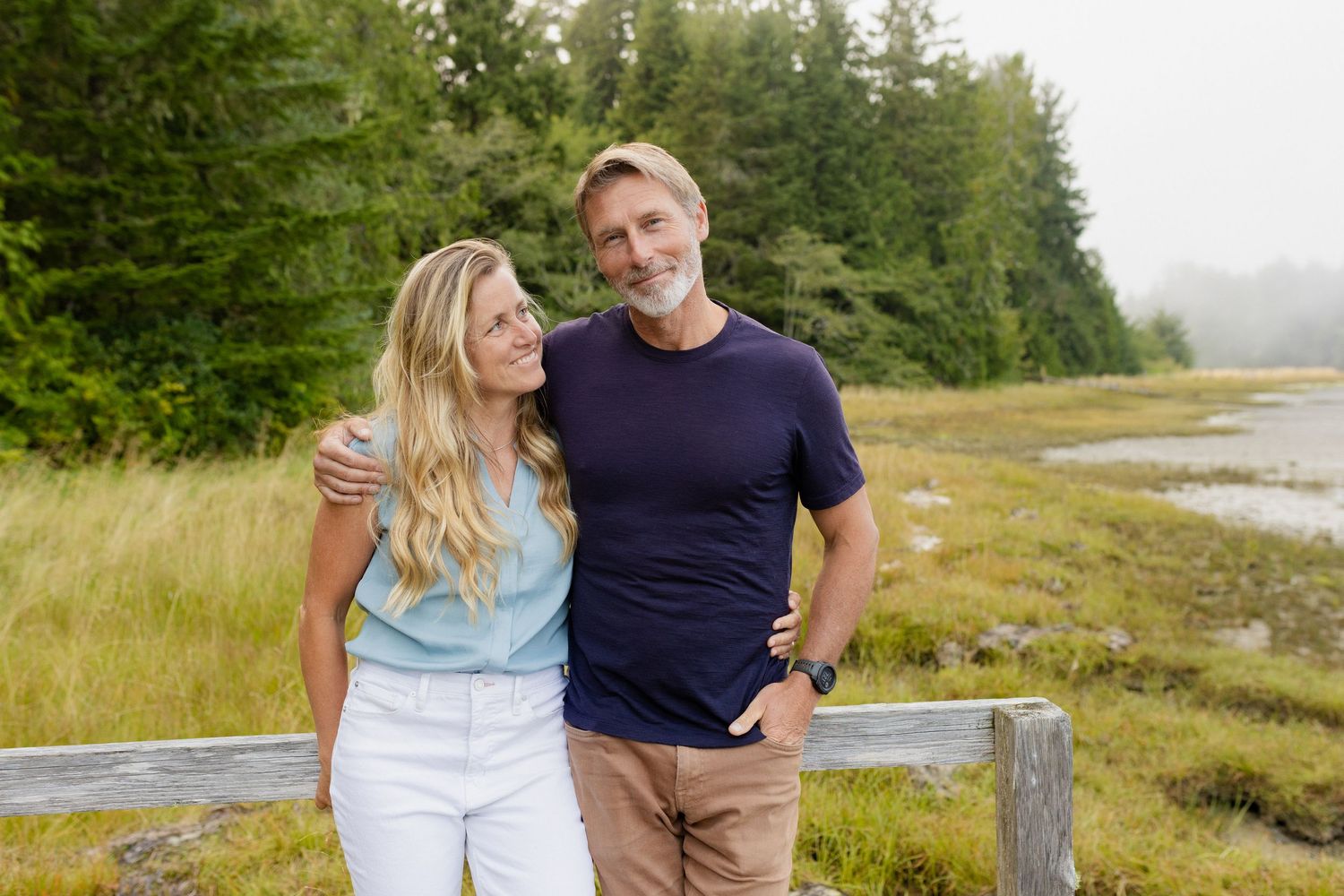 Smiling middle-aged couple embracing outdoors near wooden fence with green trees and misty field.