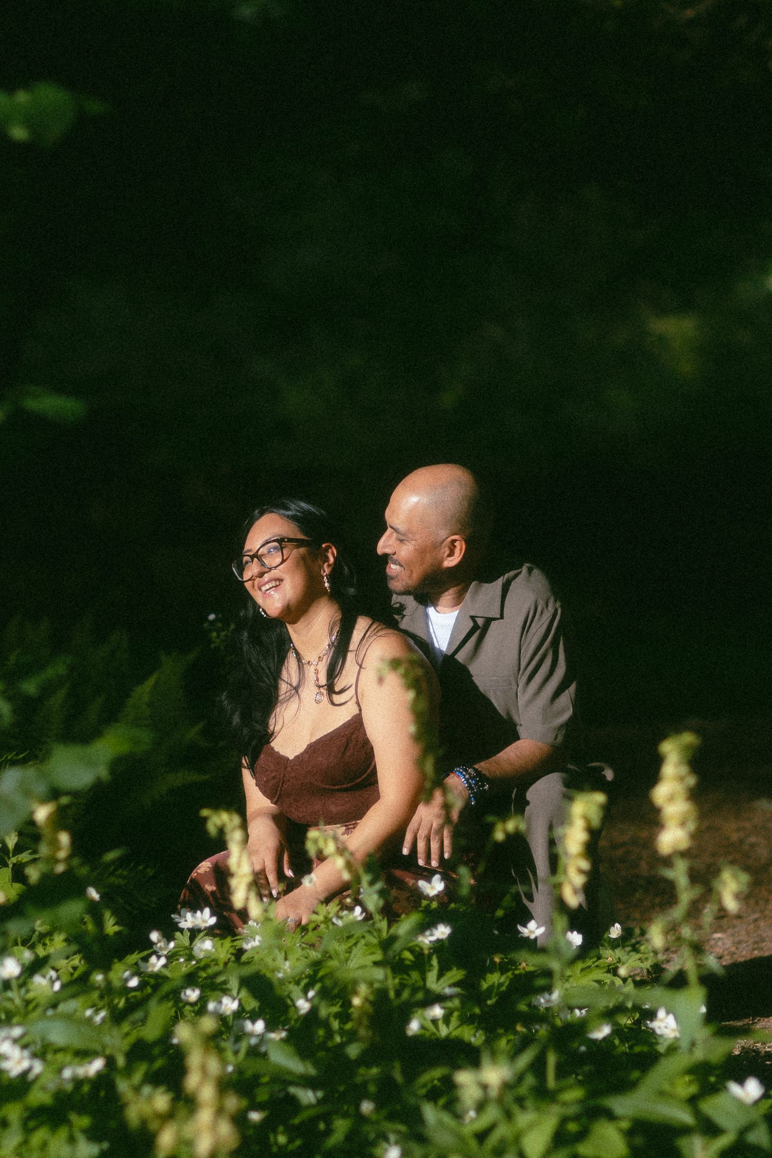A couple sits among wildflowers in a lush green forest, the woman smiling warmly while the man looks on beside her.