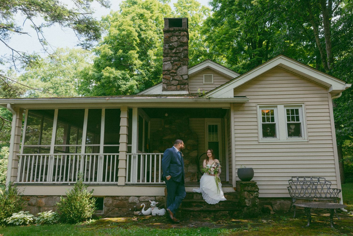 Bride and groom stand before a charming cottage with stone chimney and wraparound porch surrounded by lush trees.