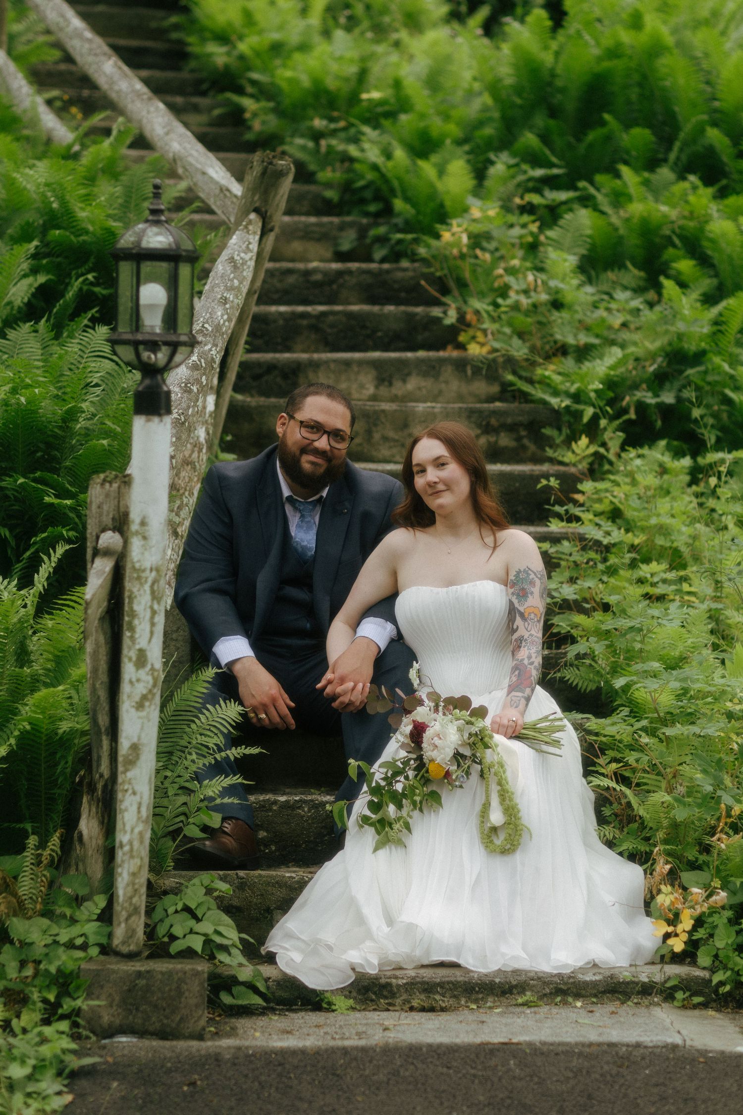 A bride and groom sit together on stone steps surrounded by lush green ferns, holding hands at an outdoor wedding.