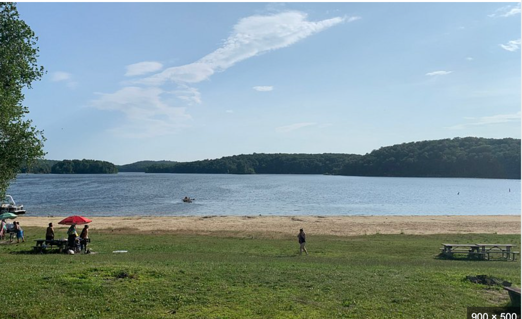 Sandy beach on a calm lake with green hills, blue sky, and visitors relaxing under umbrellas on a sunny day.